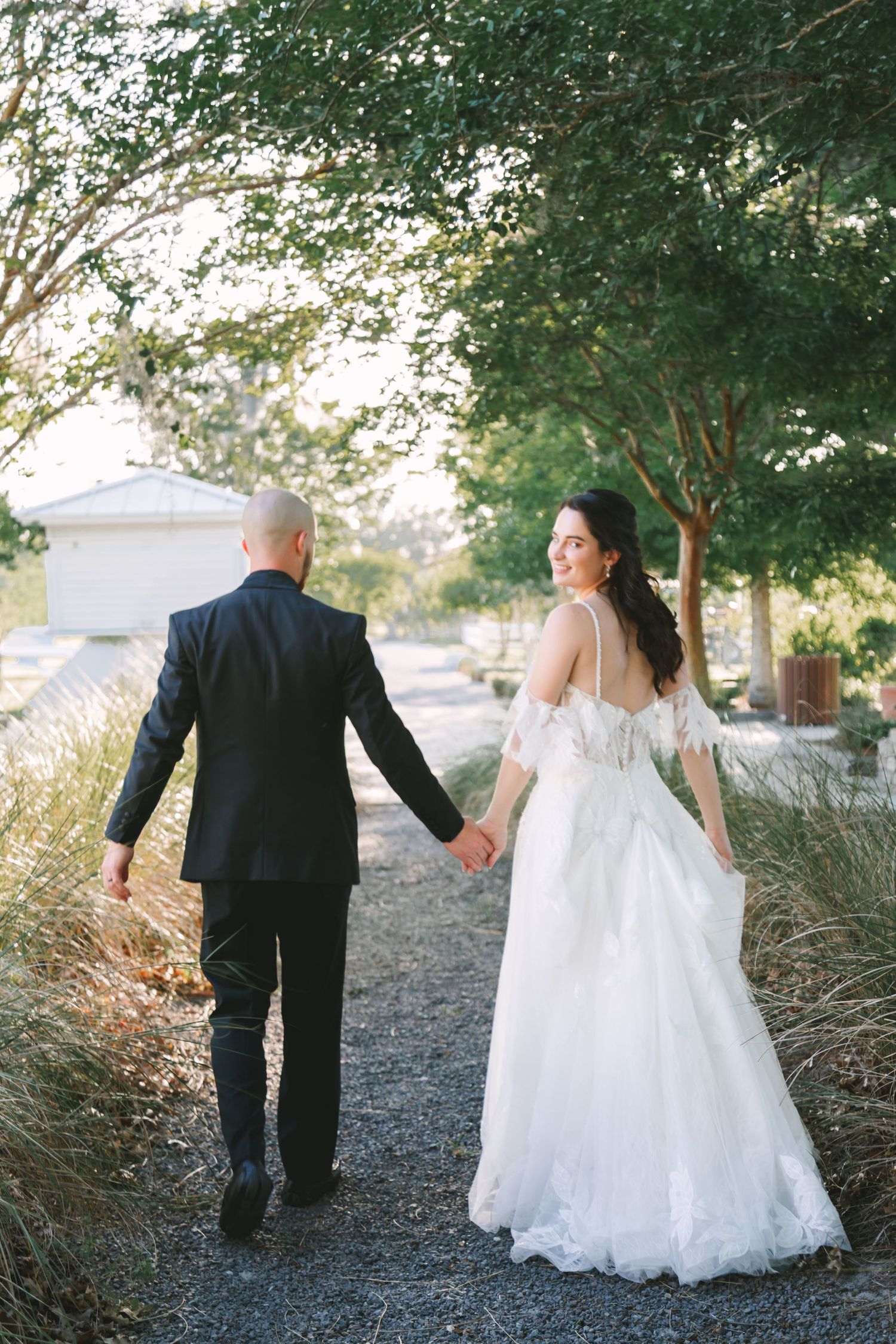 Bride in lace gown smiles back while holding grooms hand walking along garden path at sunset.
