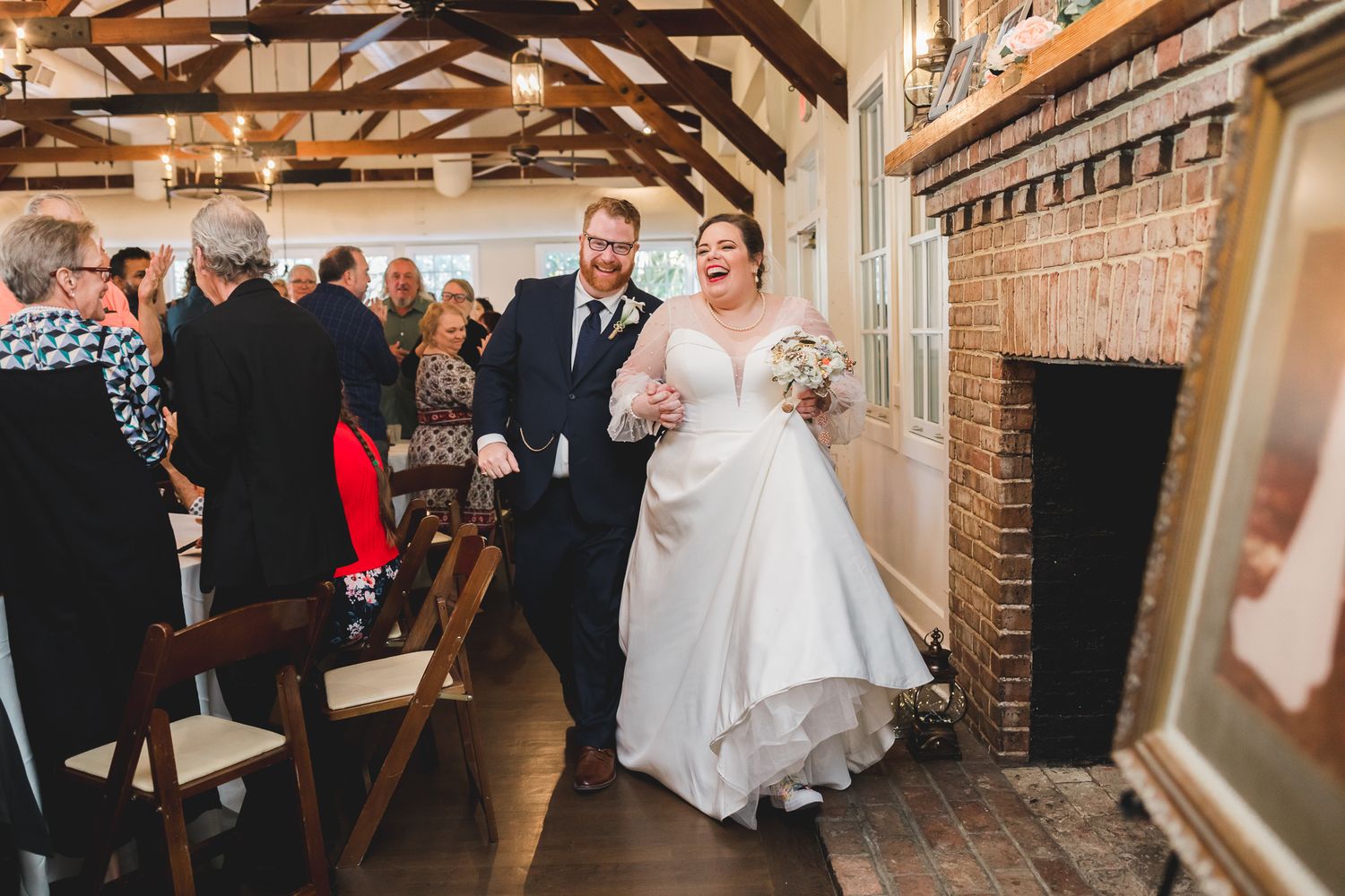 Laughing newlyweds walk down aisle past brick fireplace at intimate indoor venue.