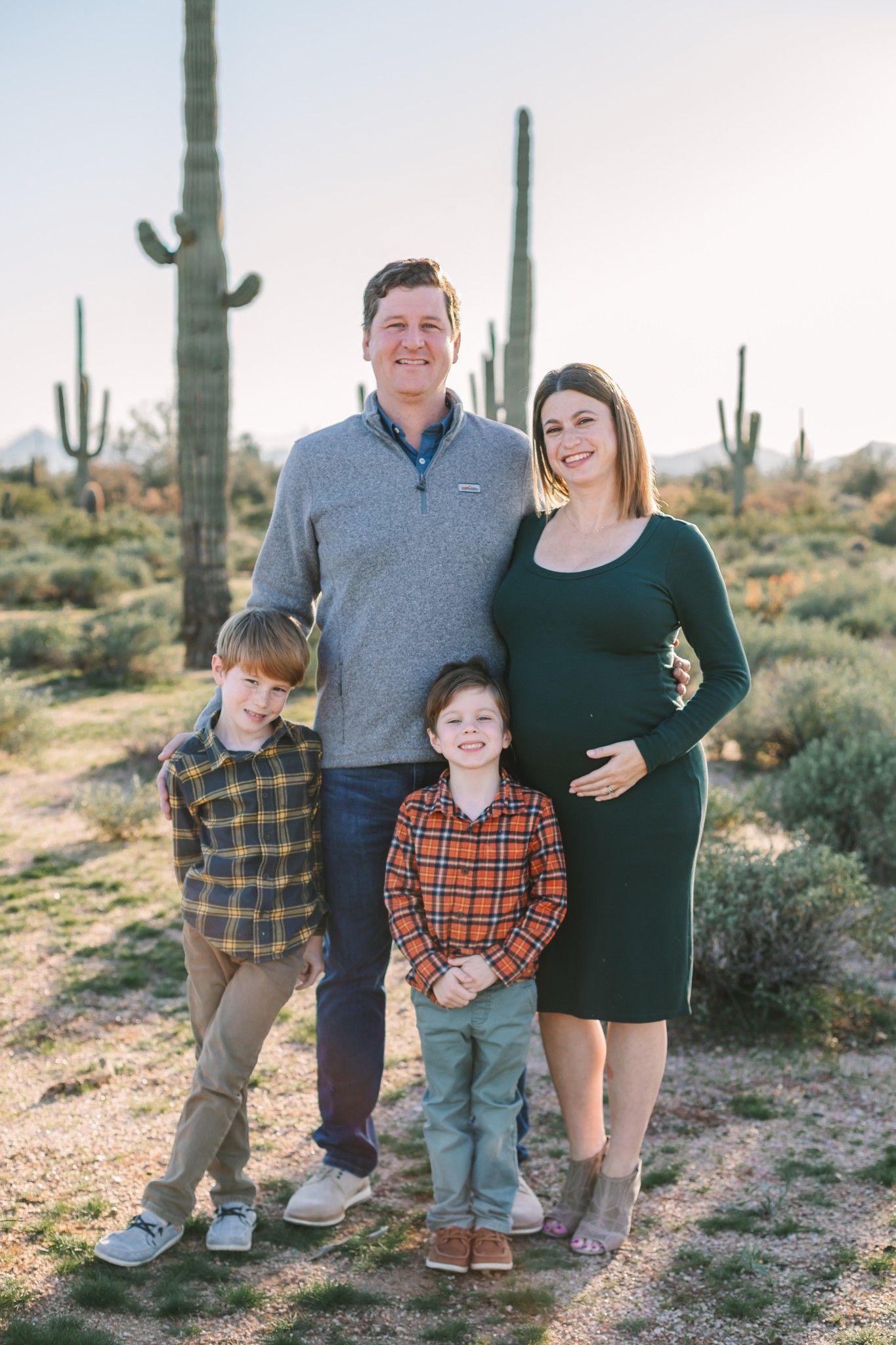 Expecting family of four poses among tall saguaro cacti in Arizona desert landscape.