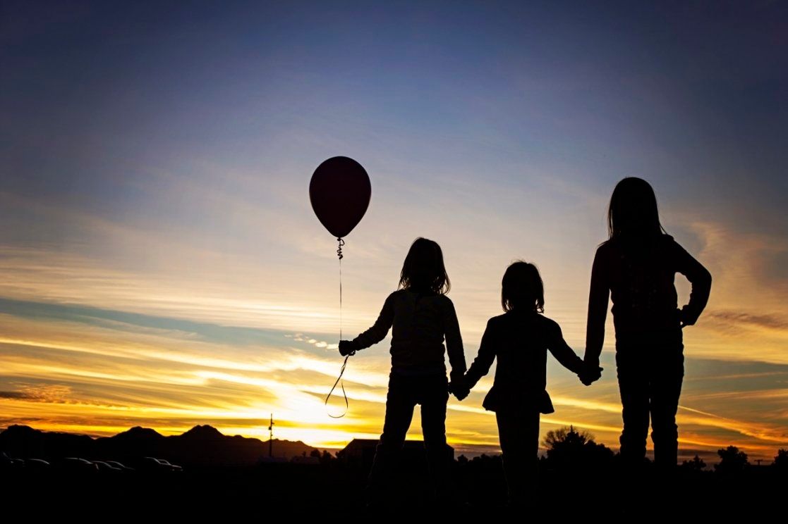 Silhouette of three girls holding hands at sunset, one holding a red balloon against colorful sky.