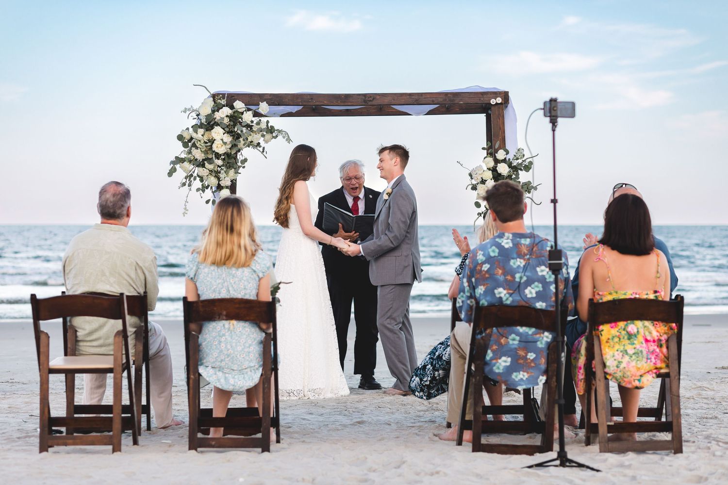 Intimate beach wedding ceremony with couple exchanging vows under a wooden floral arch at the ocean's edge.