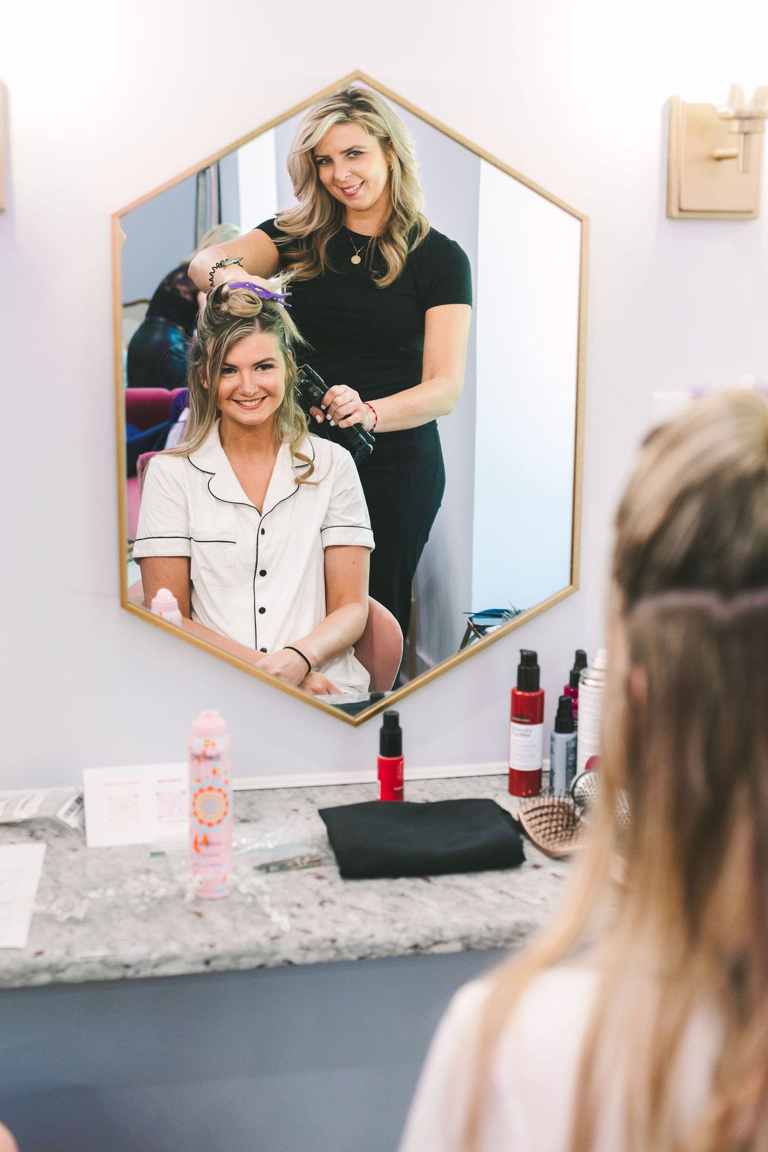 Hair stylist curling bride's hair reflected in hexagonal mirror during wedding day preparations.