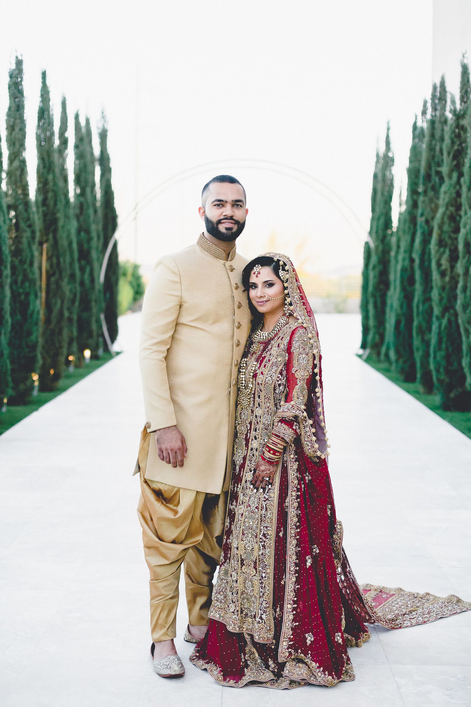 South Asian couple in traditional wedding attire posing outdoors among cypress trees.