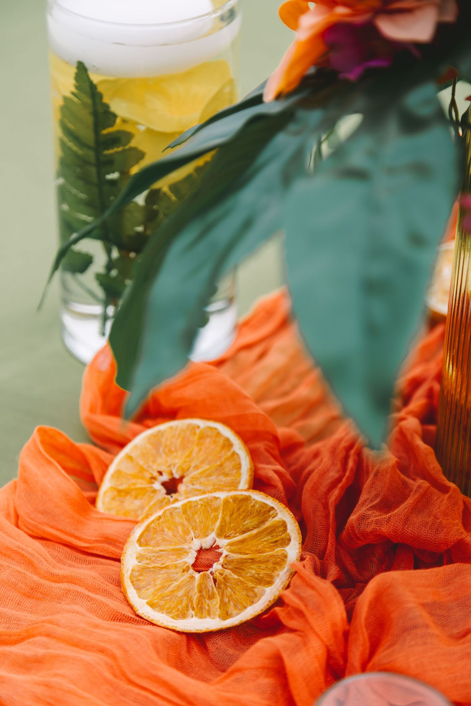 Dried orange slices on orange fabric with tropical flowers and a yellow drink in background.