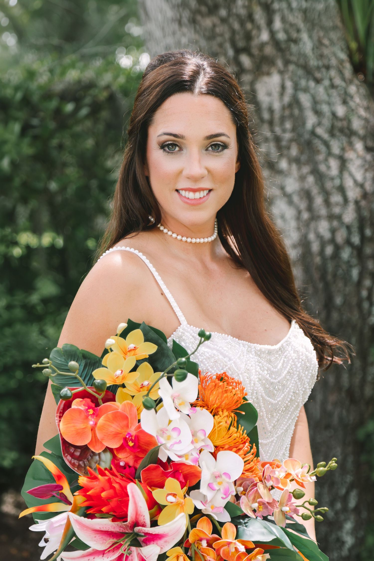Smiling bride with long brown hair holding colorful tropical bouquet with orchids and lilies outdoors.