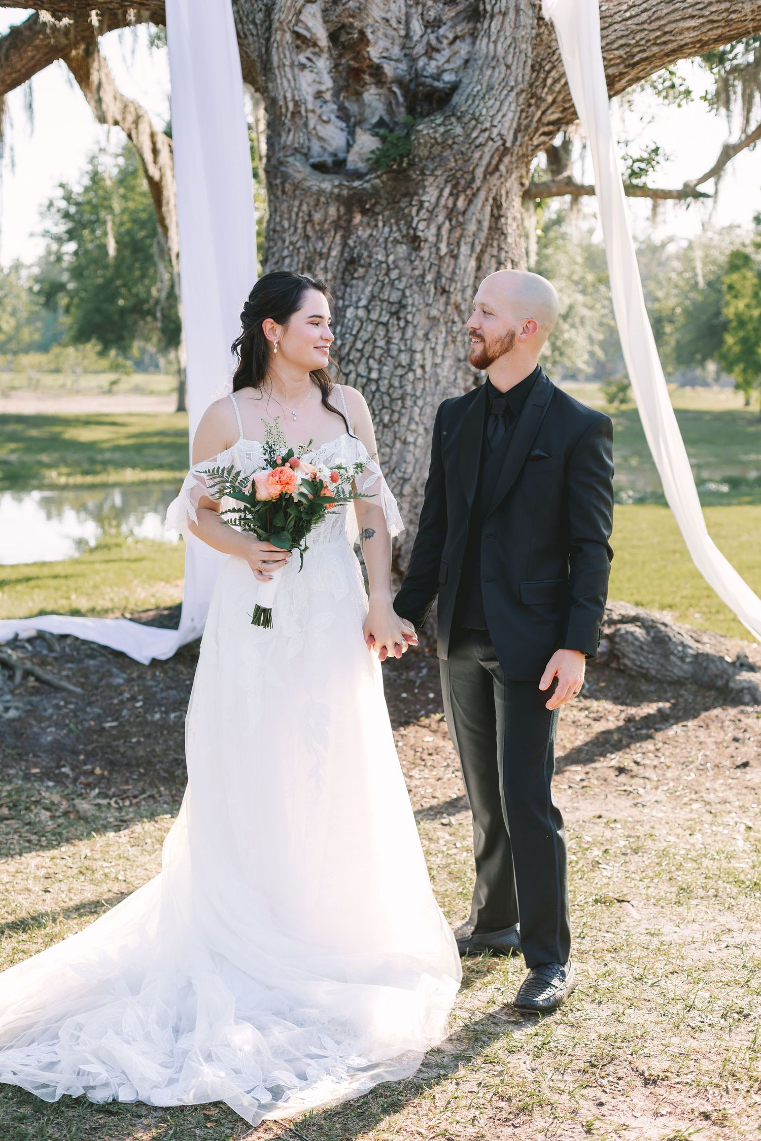 Bride in white gown holding floral bouquet walks hand-in-hand with groom in black suit outdoors near large oak tree.