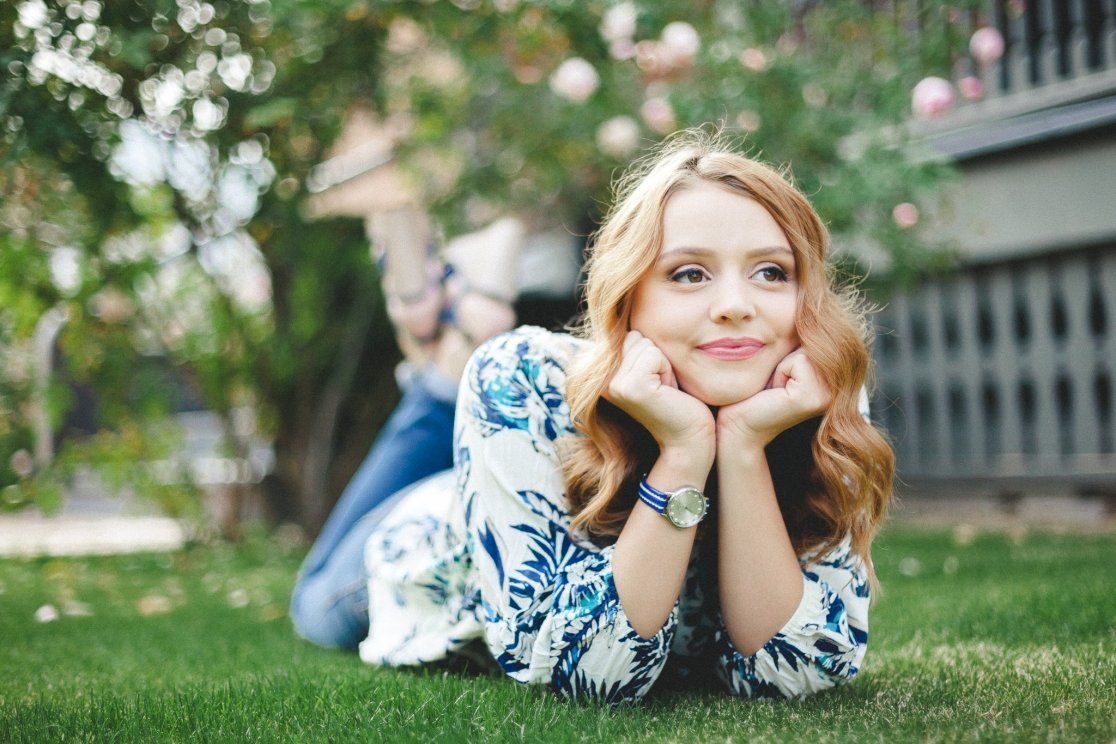 Young girl with curly blonde hair lying on green grass, smiling dreamily outdoors.