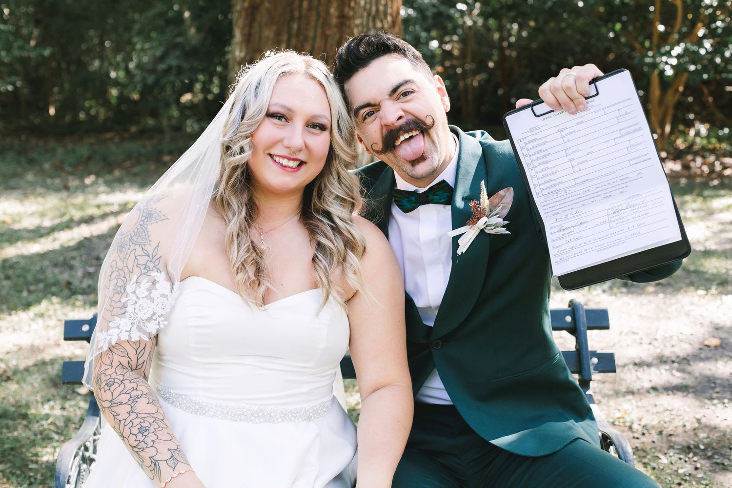 Playful bride and groom stick out tongues while holding signed marriage certificate on park bench.