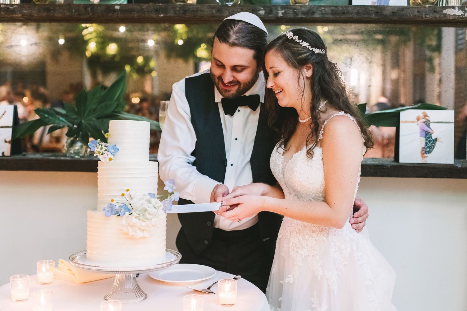 Smiling bride and groom cutting their elegant white tiered wedding cake during their indoor reception celebration.