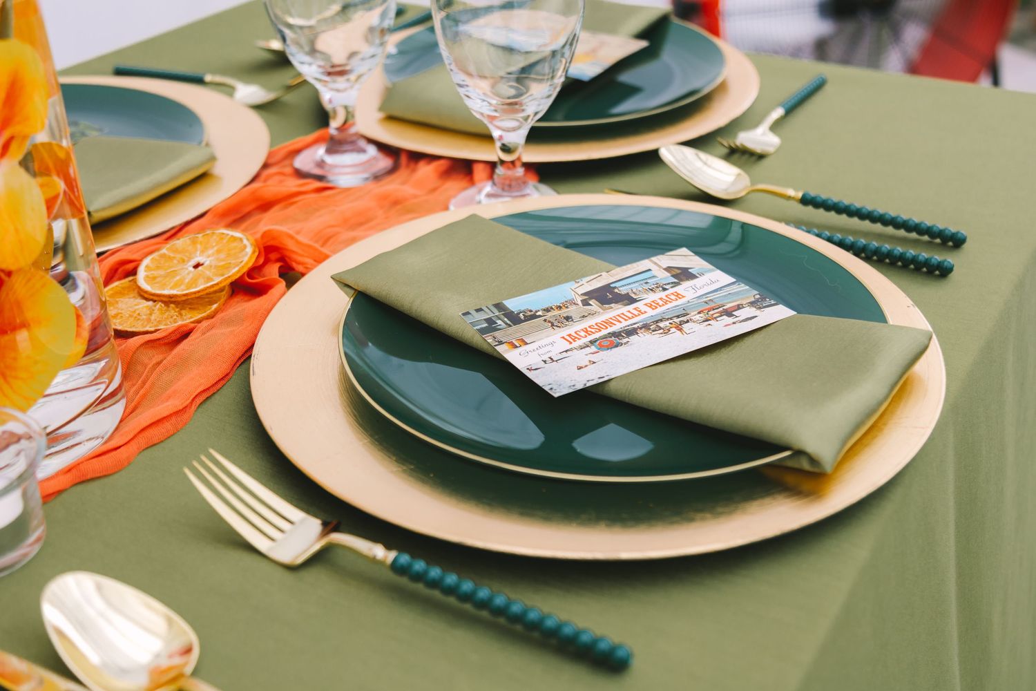 Elegant wedding table setting with green napkin, teal flatware, orange runner, and dried orange slice decor.