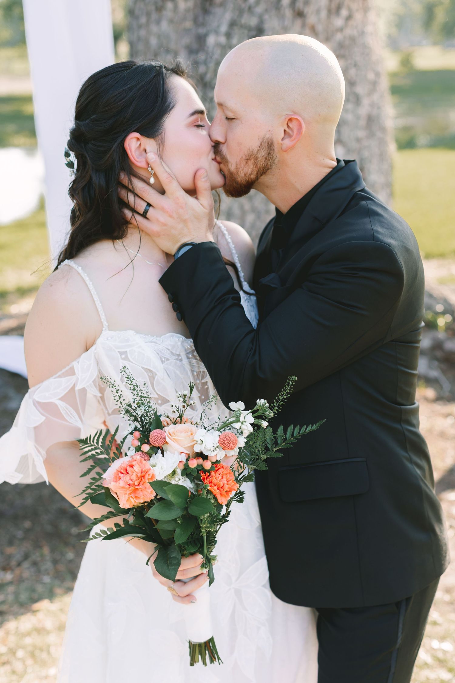 Bride and groom share a romantic first kiss, bride holding a coral and white bouquet at outdoor wedding.