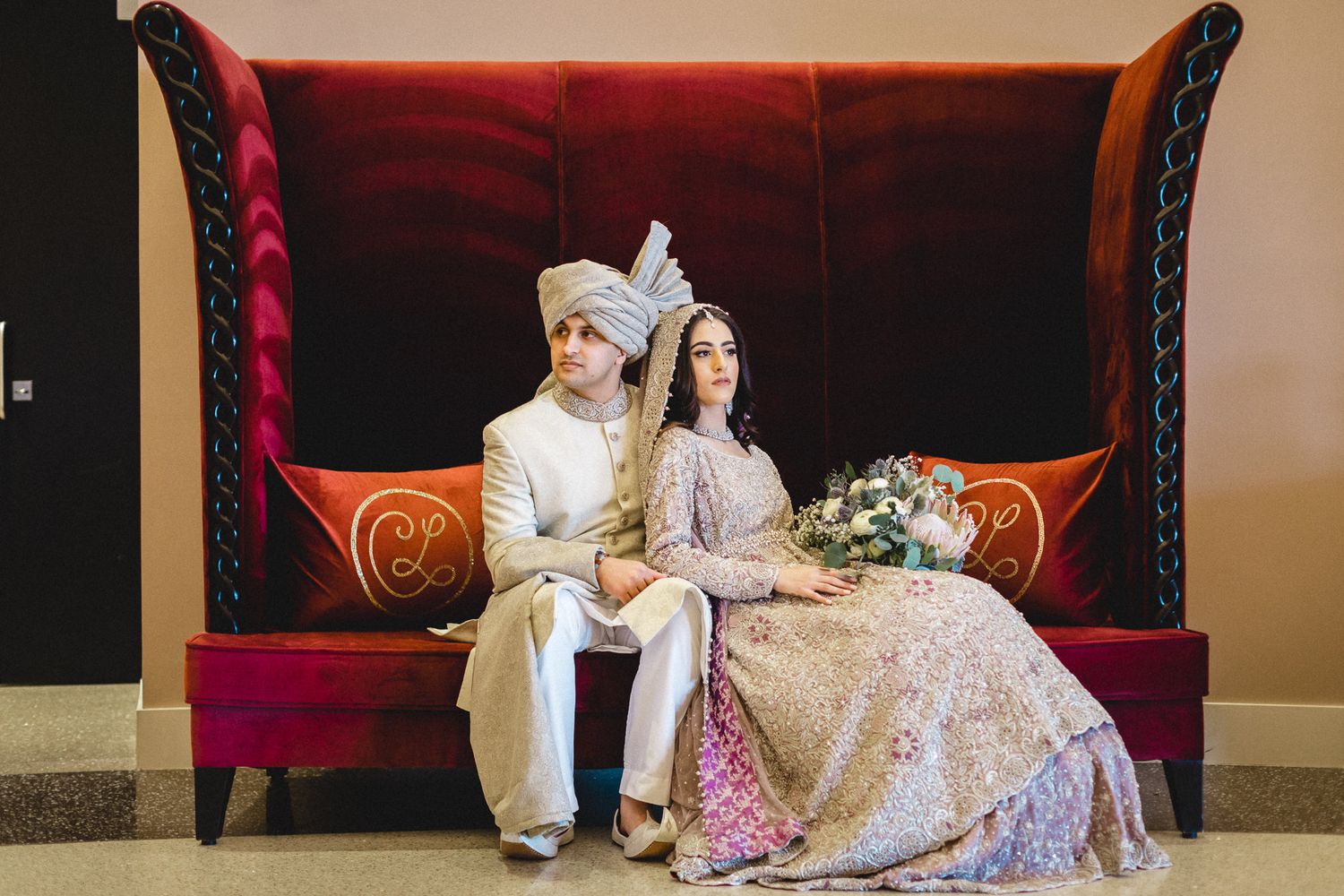 South Asian wedding couple in traditional attire sitting on ornate red velvet sofa with bouquet.