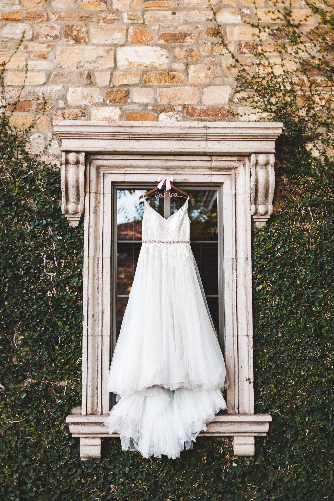 Elegant white wedding dress hanging on ornate stone window frame covered in lush green ivy.