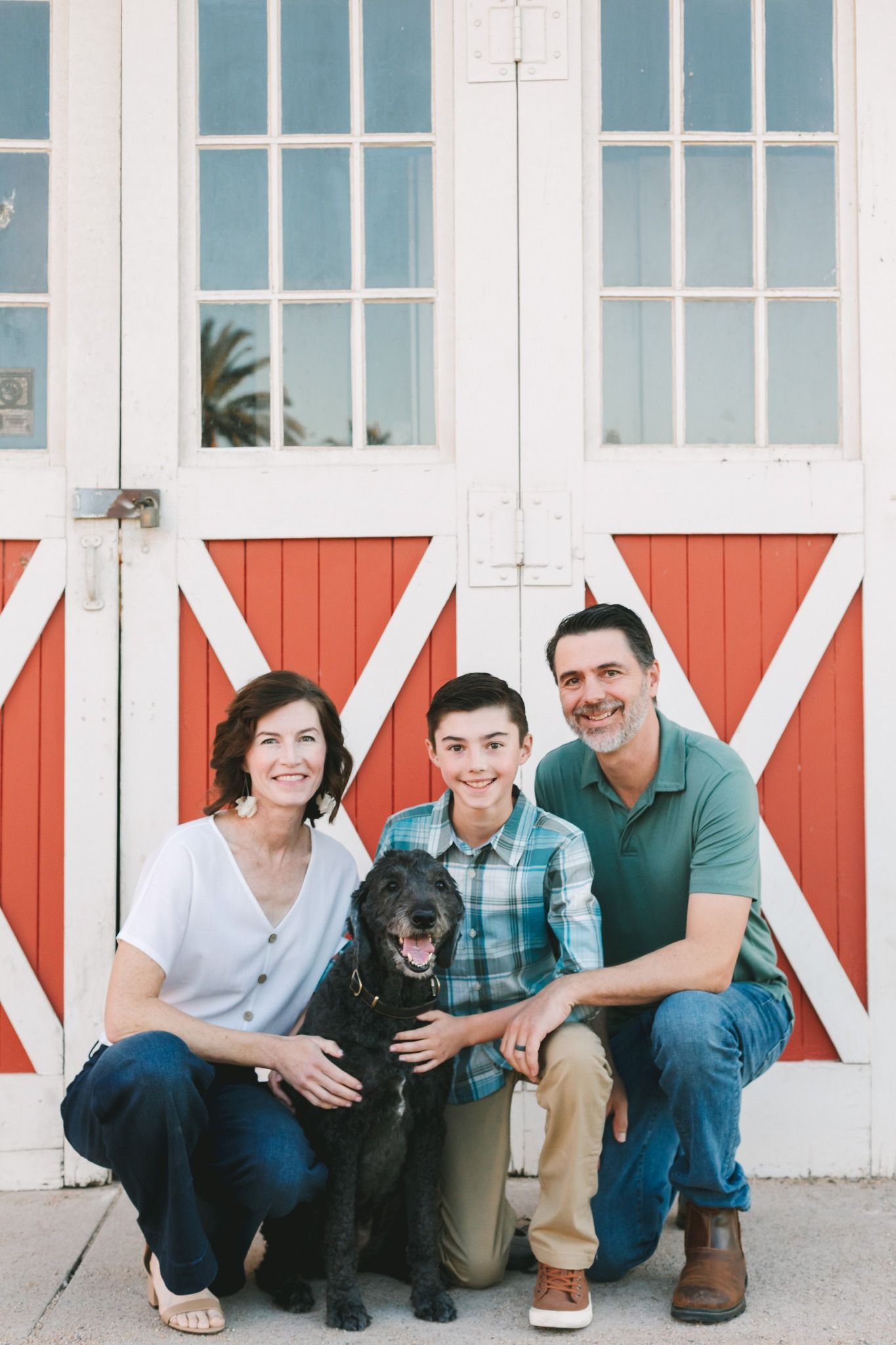 Smiling family with teenage son and black dog pose before red barn doors.