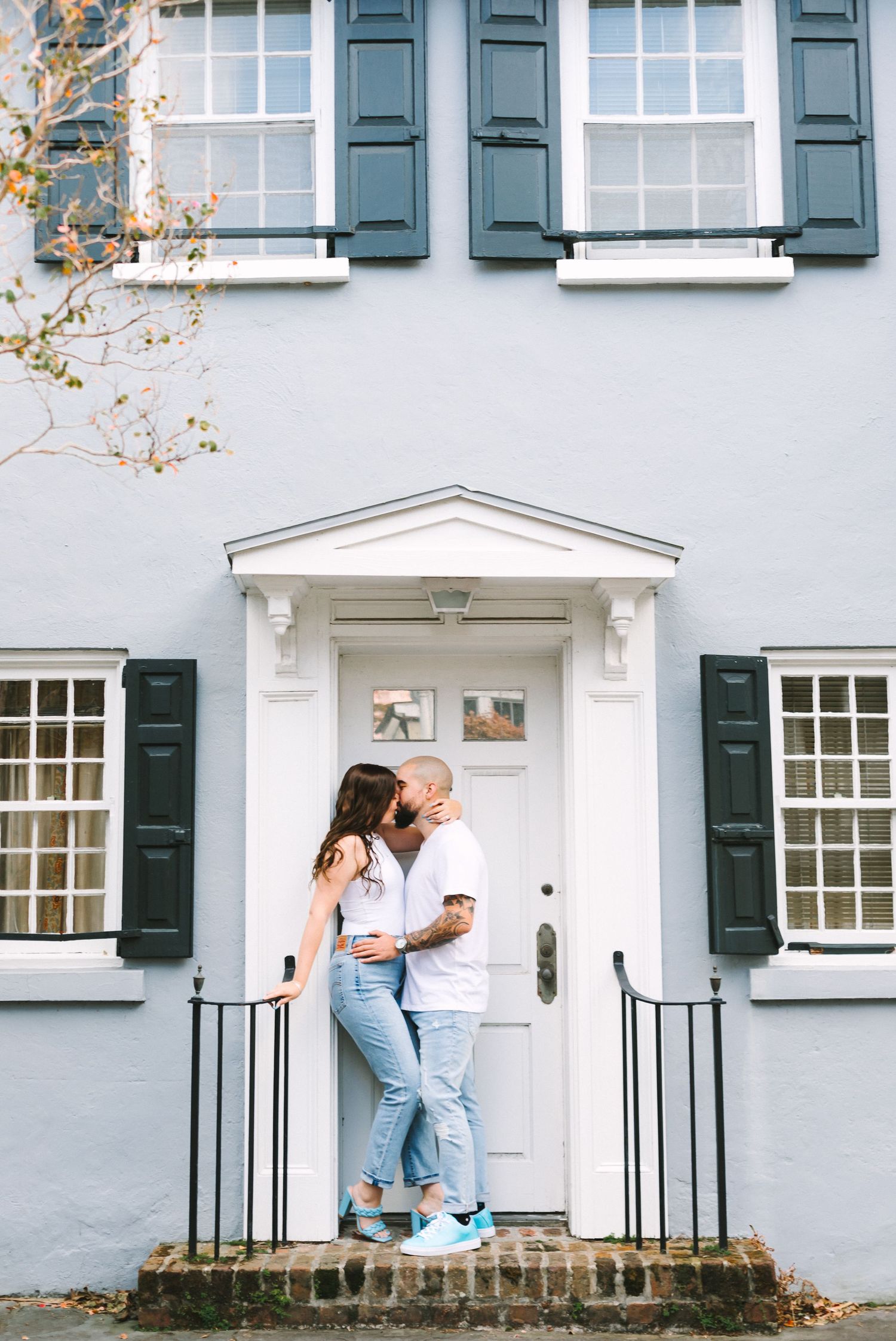 Couple sharing a kiss on the steps of a charming blue colonial home with dark shutters.