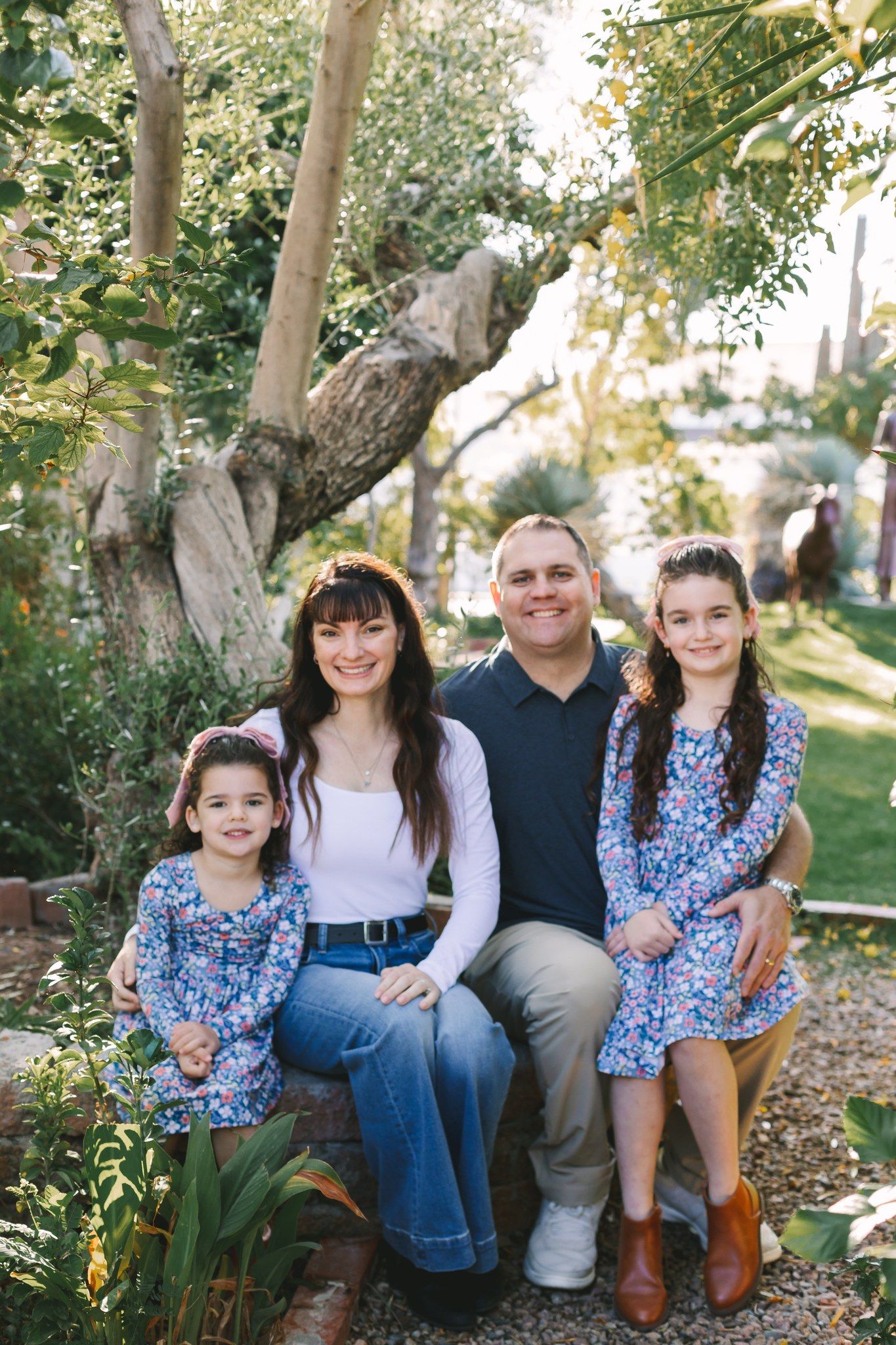 Family of four sitting together outdoors in a lush garden setting, two daughters in matching floral dresses.