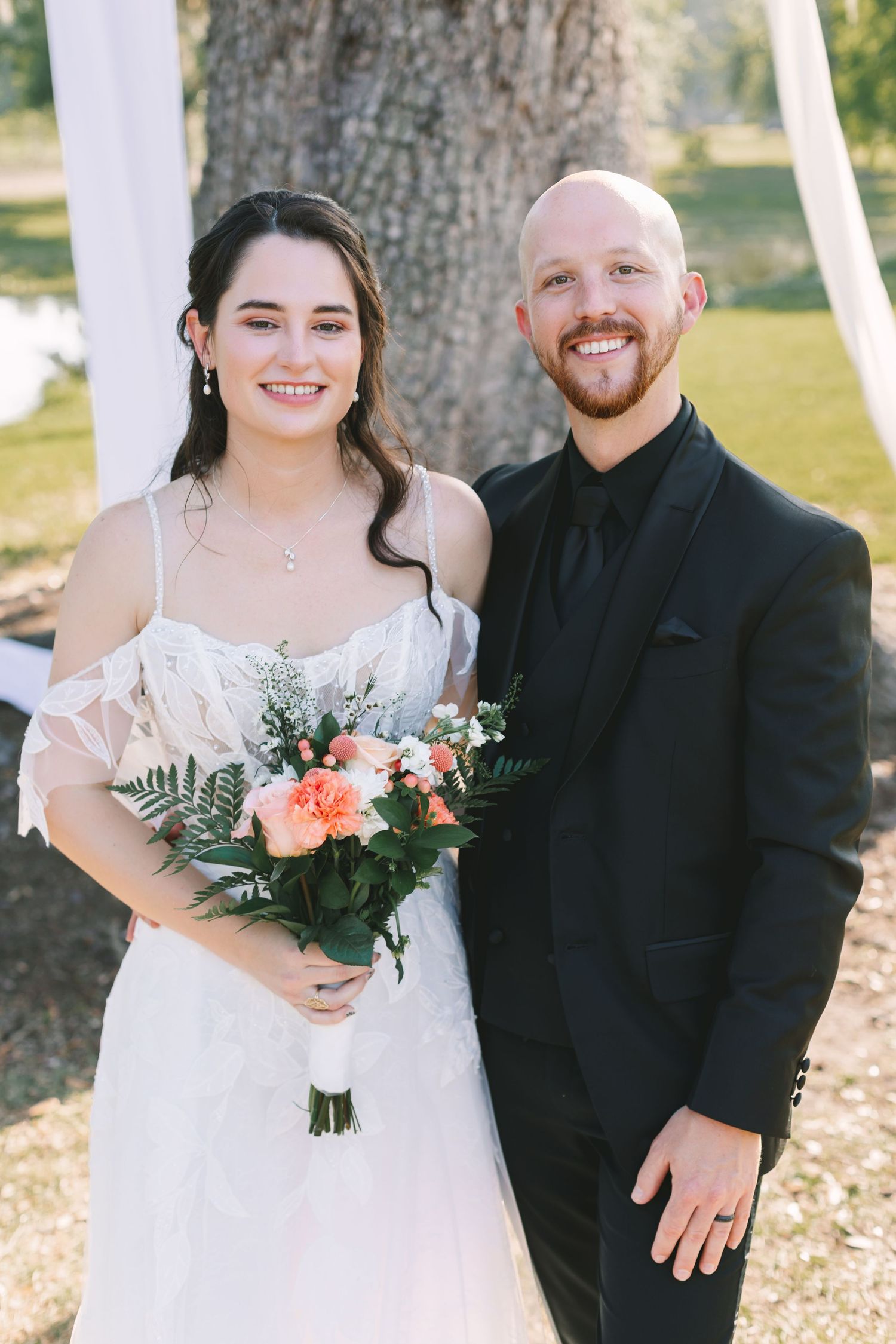 Smiling bride holding peach bouquet stands with bald groom in black suit outdoors at wedding ceremony.