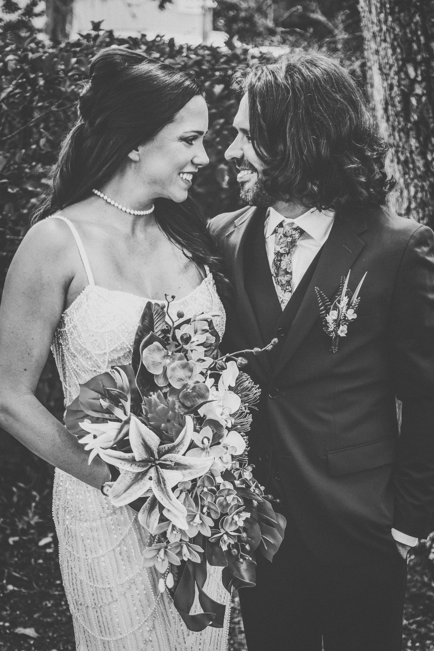 Black and white photo of a smiling bride holding a bouquet and groom in a suit gazing at each other on their wedding day.