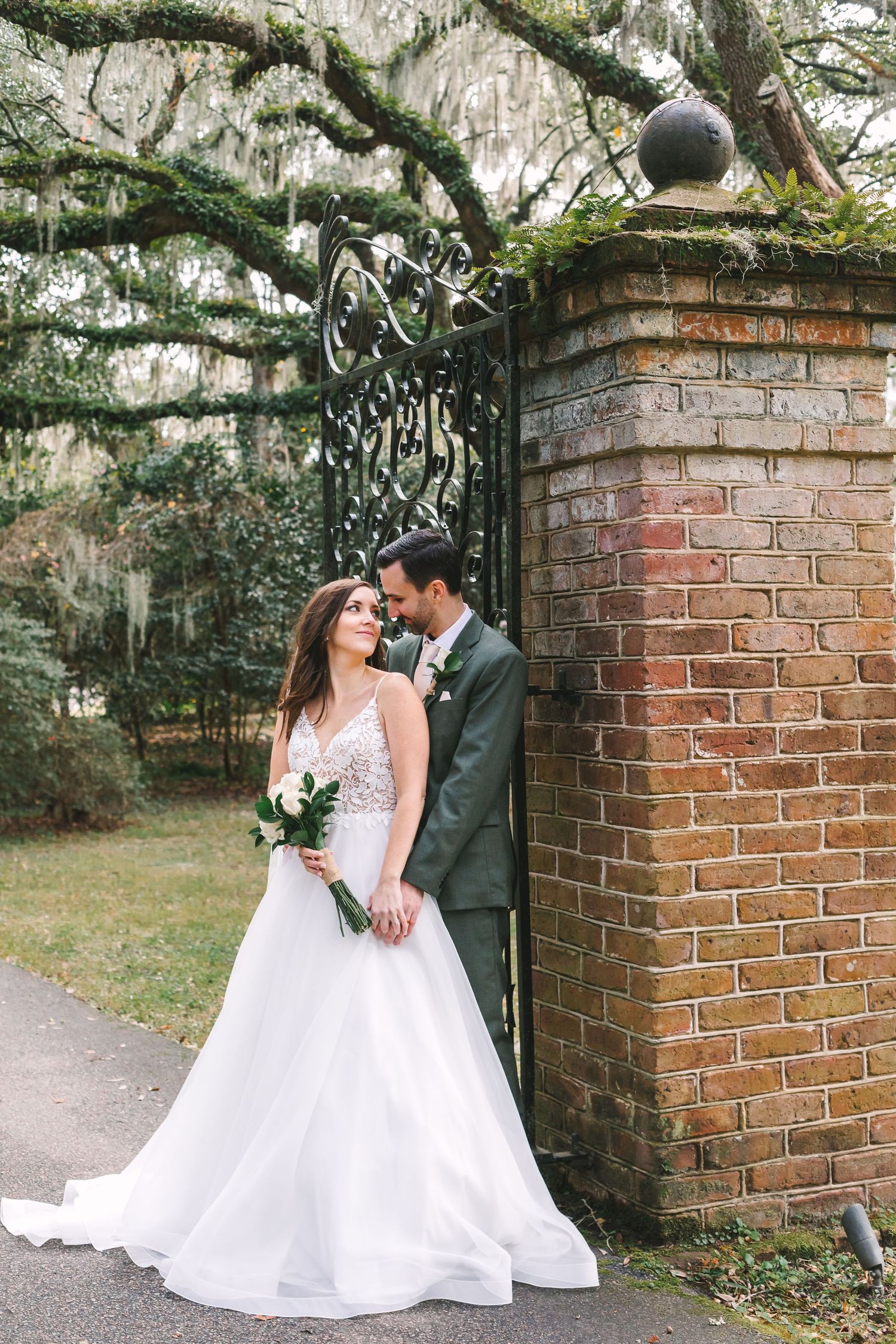 Bride and groom embrace by ornate iron gate and brick pillar, surrounded by moss-draped oak trees on their wedding day.
