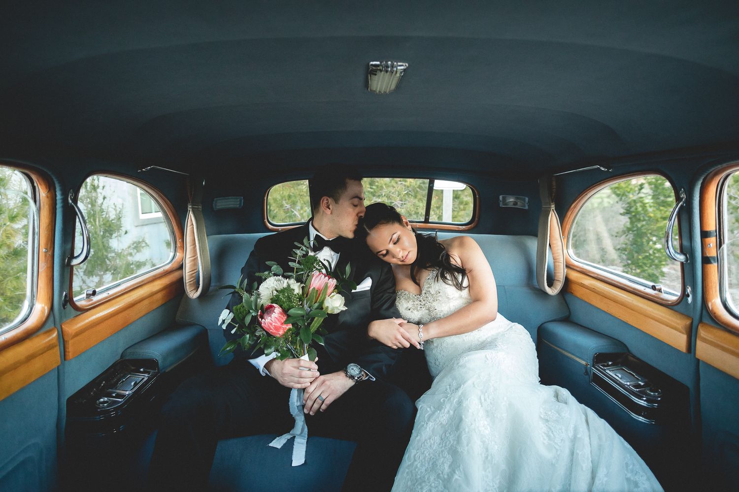 Newlywed couple sharing intimate moment in vintage car backseat with bridal bouquet.