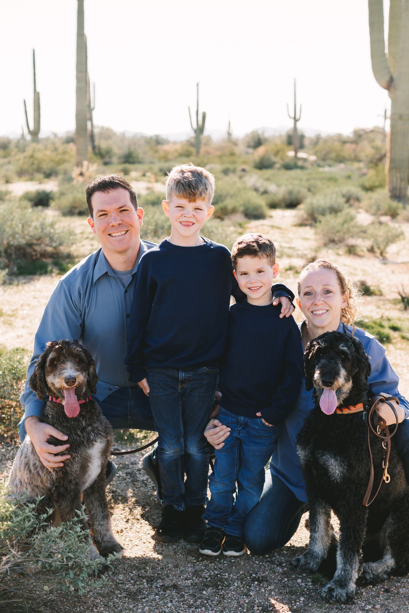 Smiling family with two dogs posing in Arizona desert with saguaro cacti background.