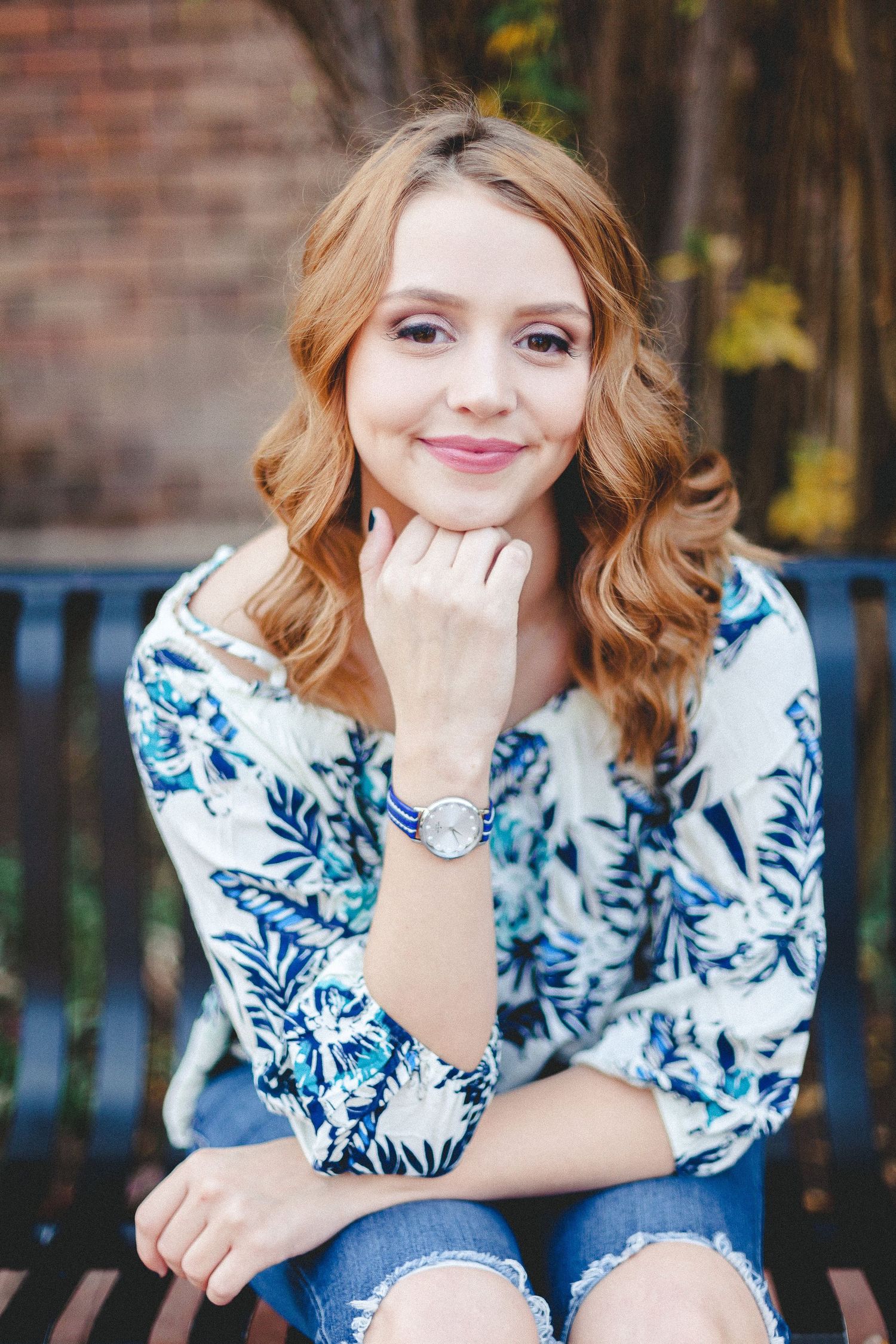Young woman with curly auburn hair smiling on bench wearing floral top and ripped jeans.
