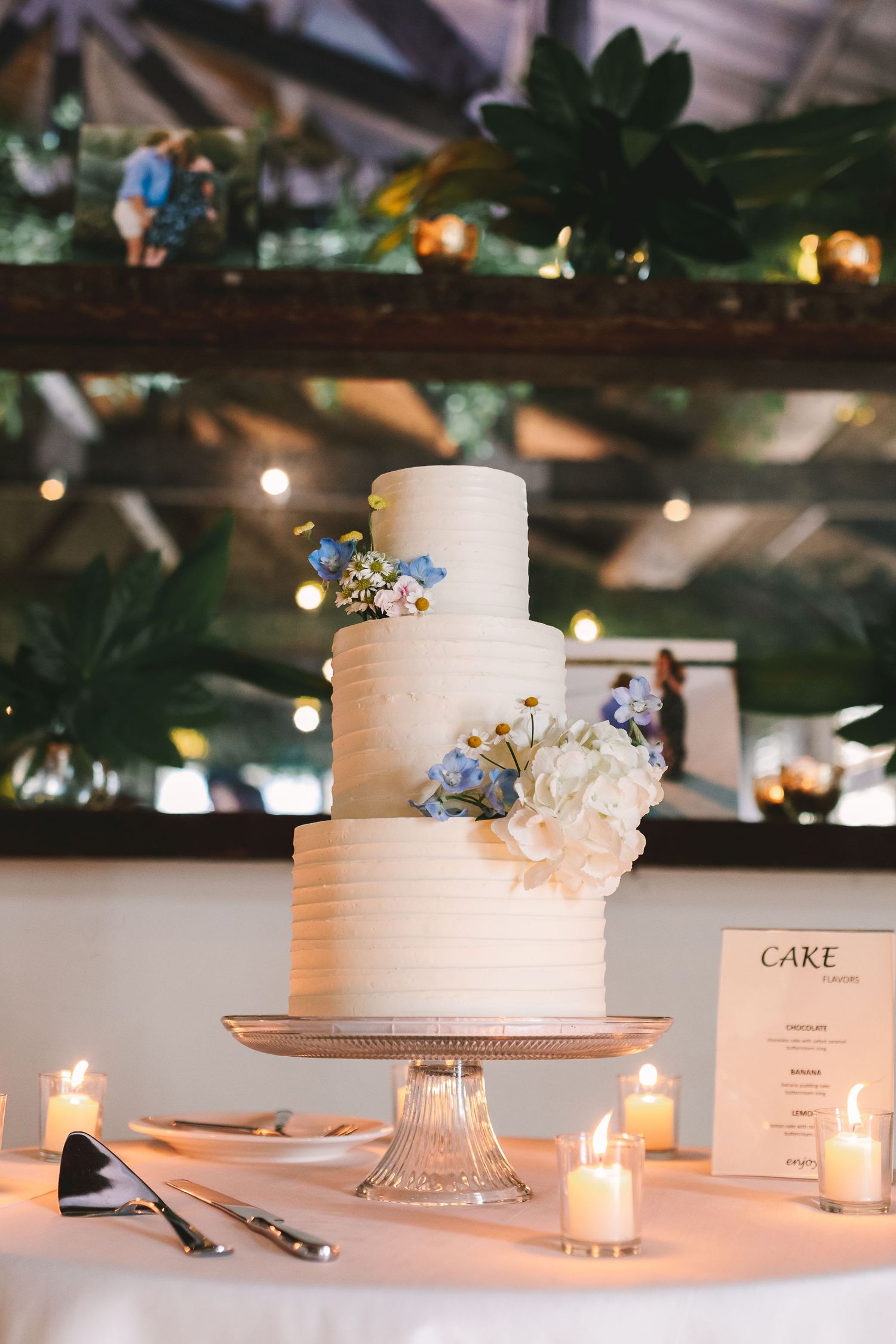 Elegant three-tier white wedding cake adorned with blue and white flowers on a silver stand surrounded by candles.