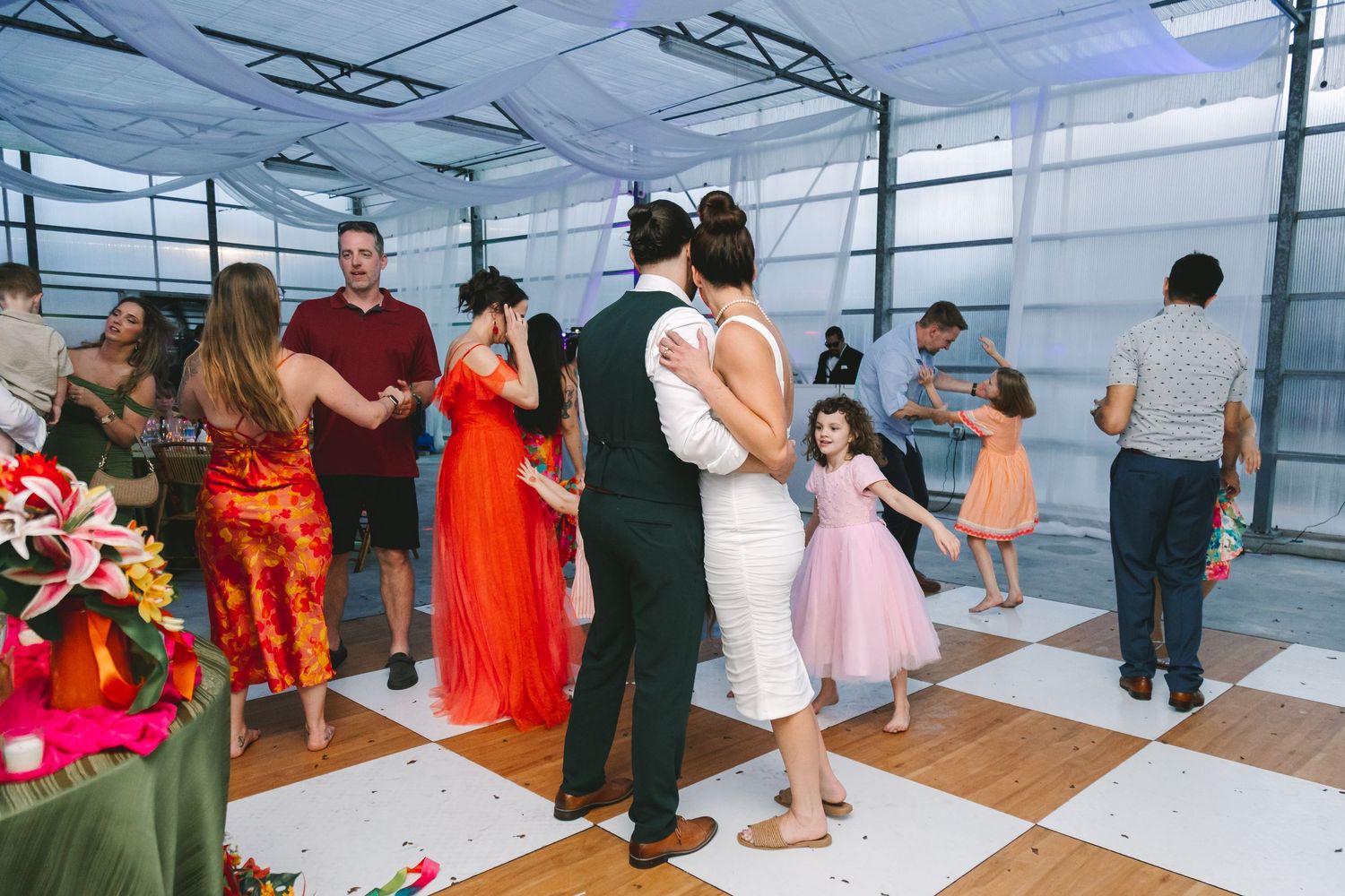 Wedding guests dancing on white checkered floor in modern venue with colorful decorations.