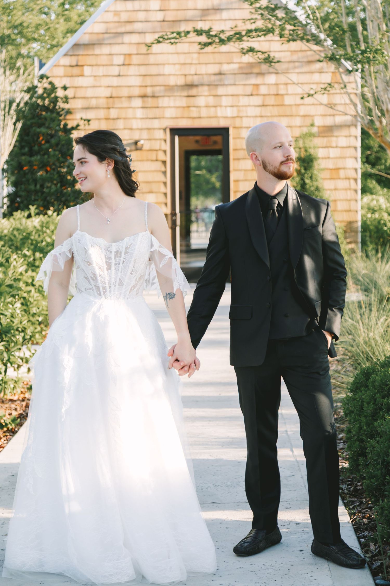 Bride in off-shoulder white gown holds hands with bald groom in black suit outdoors.