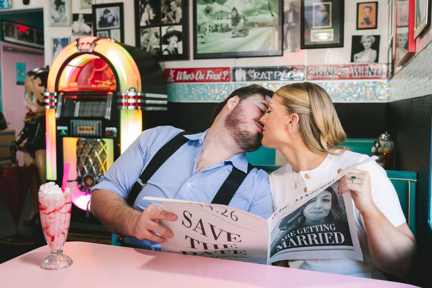 Couple kissing at retro diner booth with Save the Date sign, milkshake, and colorful jukebox in background.