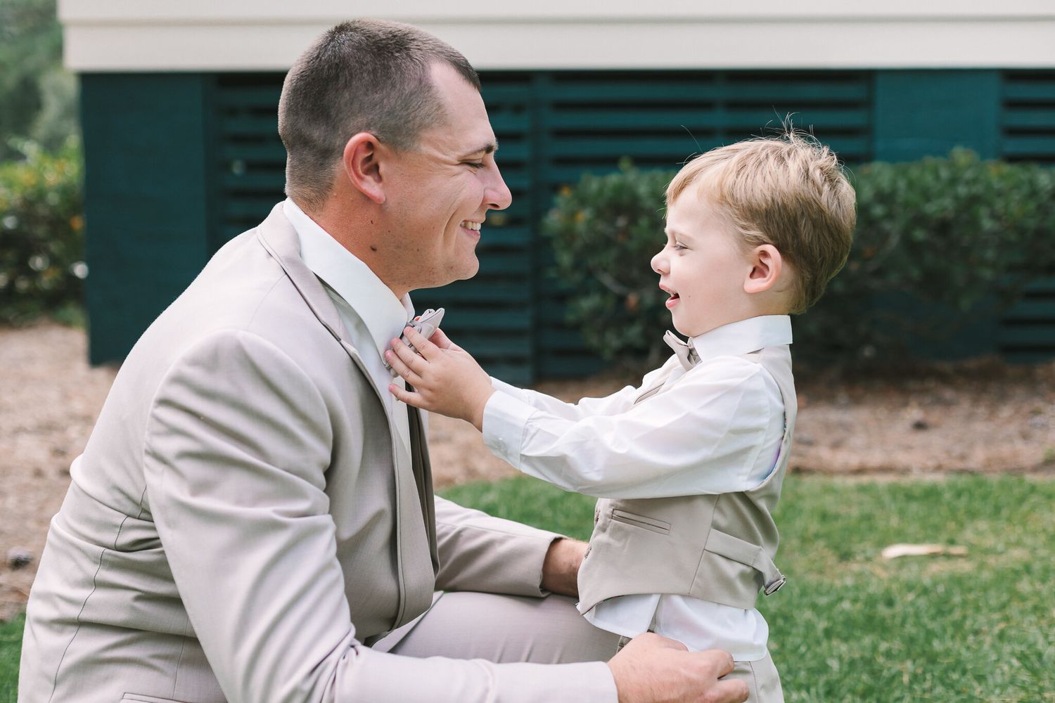 Father in tan suit smiling as young boy adjusts his tie outdoors at wedding.