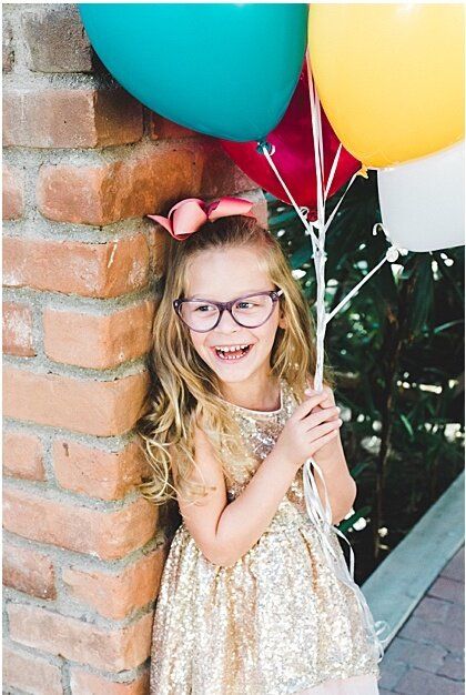 Happy young girl with glasses and pink bow holding colorful balloons in a gold sequin dress by a brick wall.