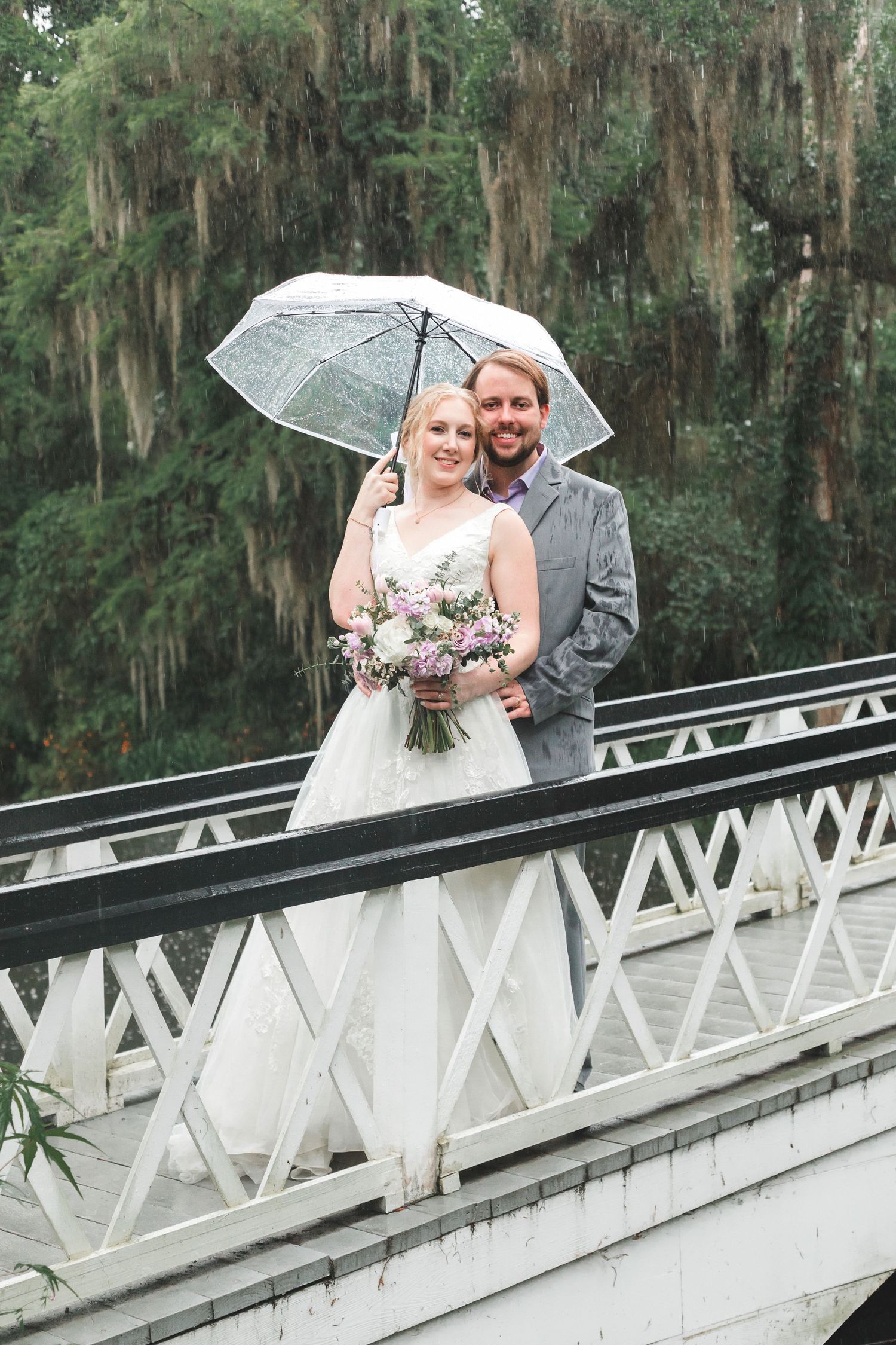 Bride and groom pose on white bridge under umbrella in rain, surrounded by Spanish moss trees.