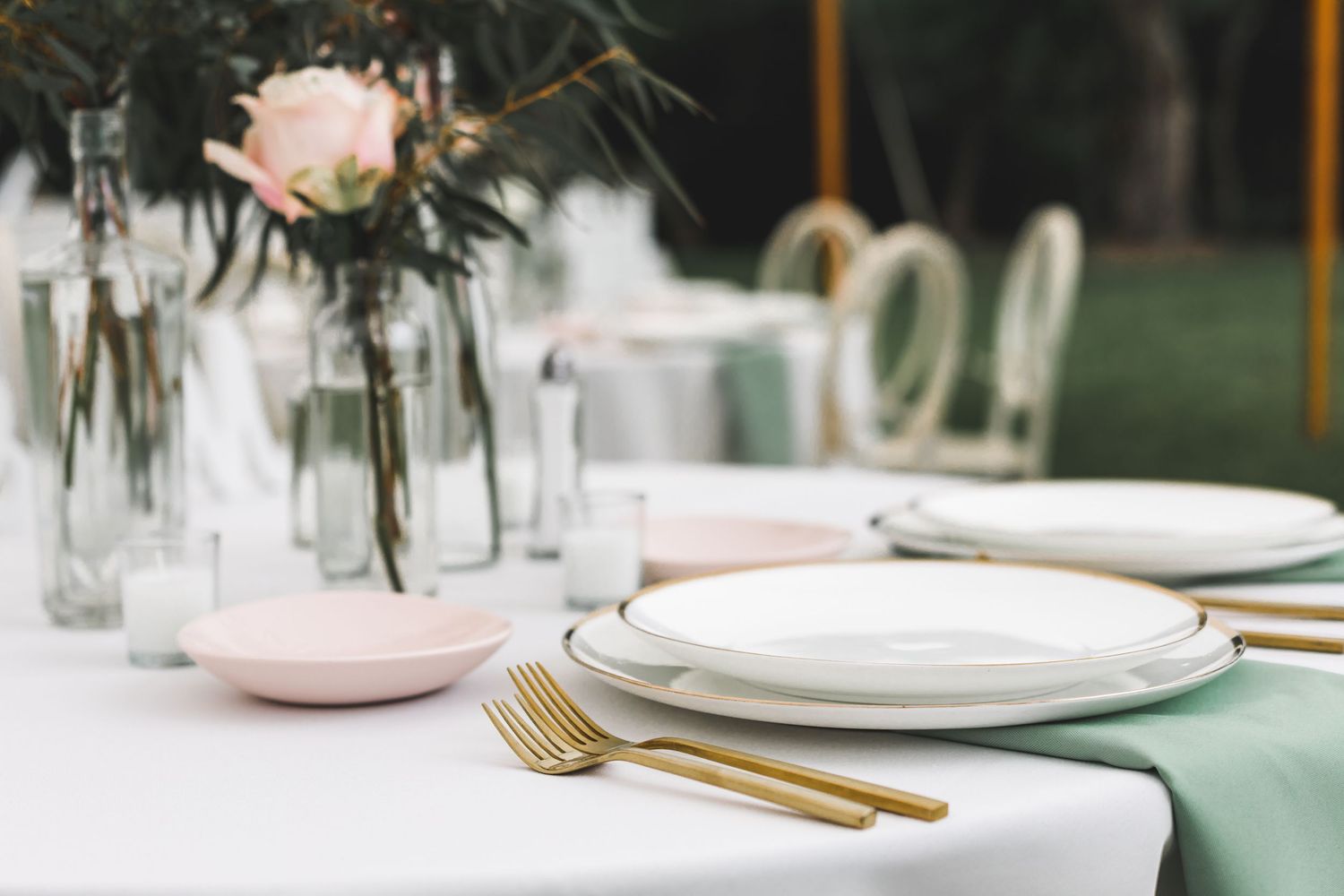 Elegant wedding table setting with gold cutlery, white plates, sage green napkins, and pink rose centerpieces.