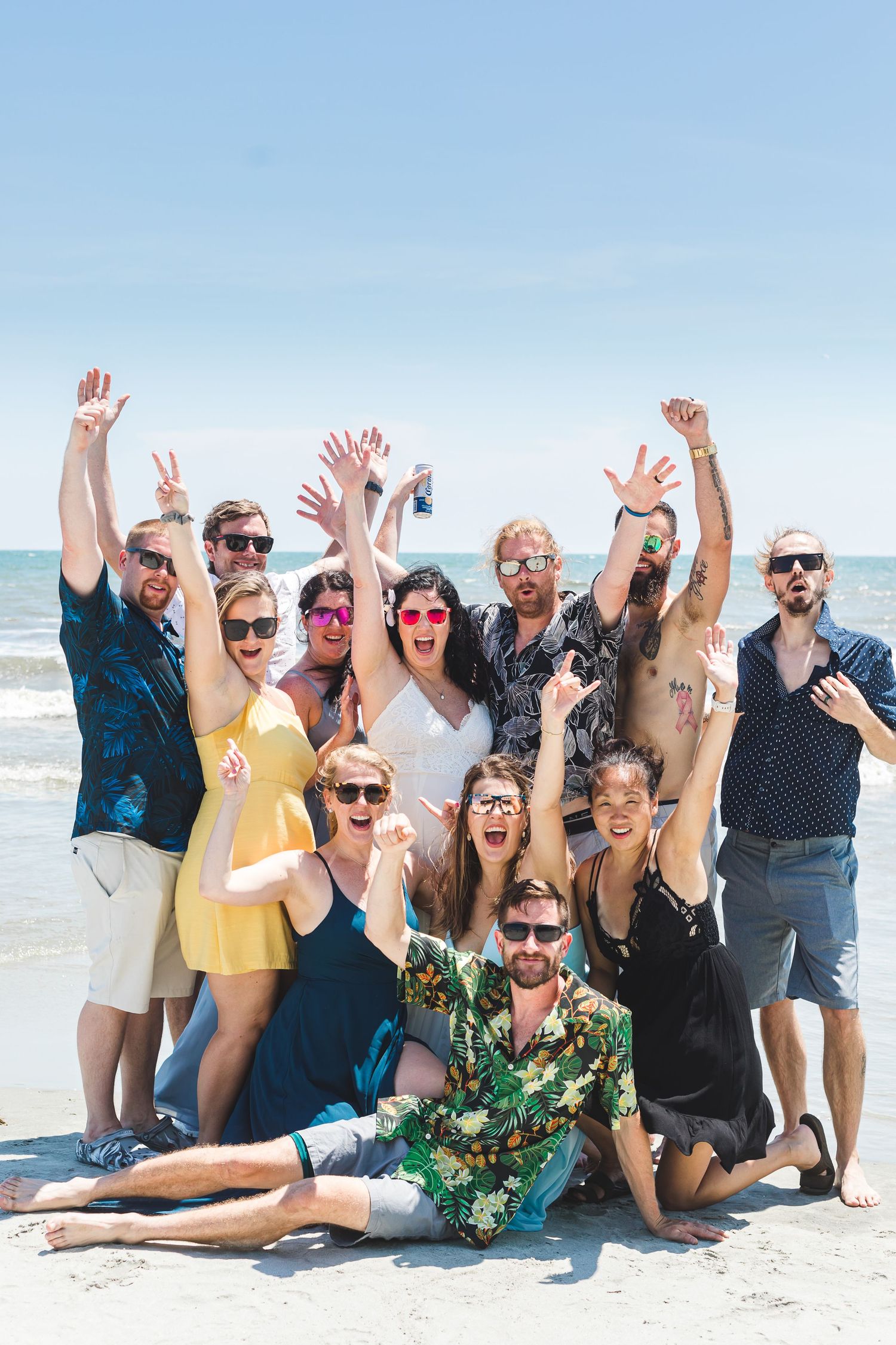 Group of wedding guests celebrating on a sunny beach with arms raised, wearing colorful summer attire.