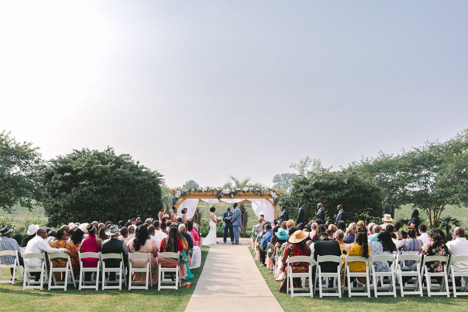Outdoor wedding ceremony with guests seated on white chairs facing a floral arch altar on green lawn.