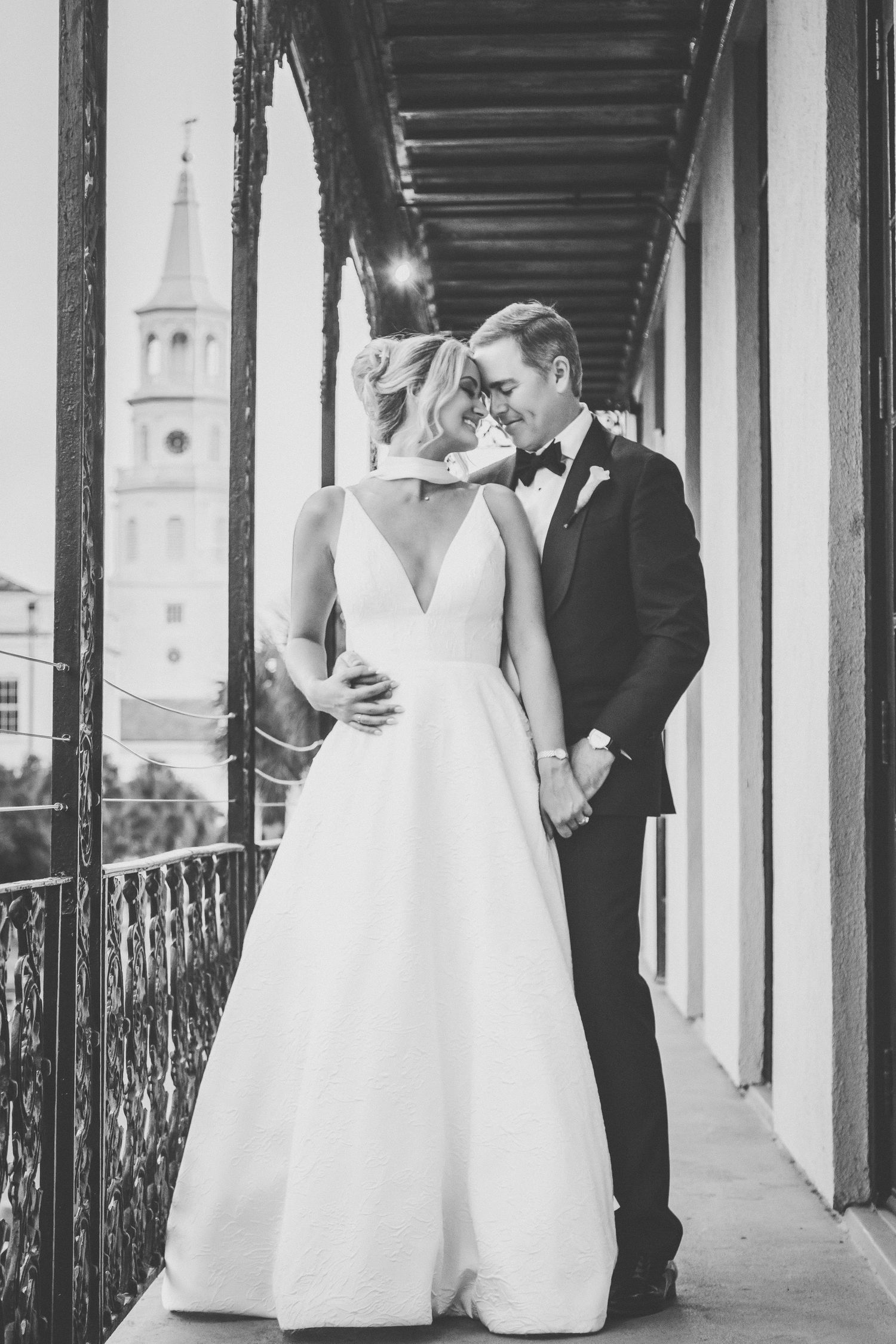 Black and white portrait of bride and groom sharing an intimate moment on a historic balcony with church steeple.