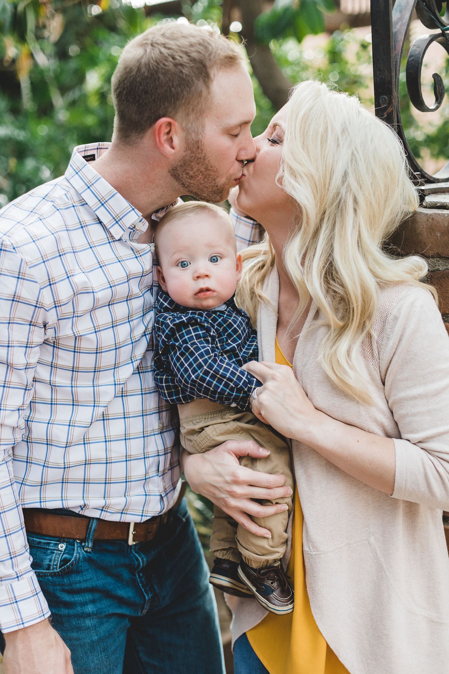 Couple sharing a kiss while holding their wide-eyed baby boy outdoors near iron gate.