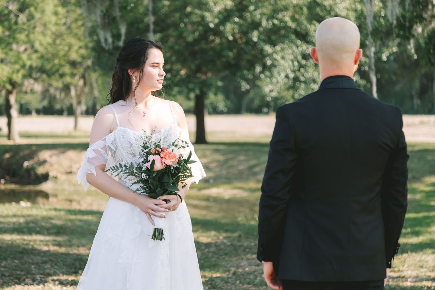 Bride in white off-shoulder gown holds peach bouquet facing groom in black suit outdoors.
