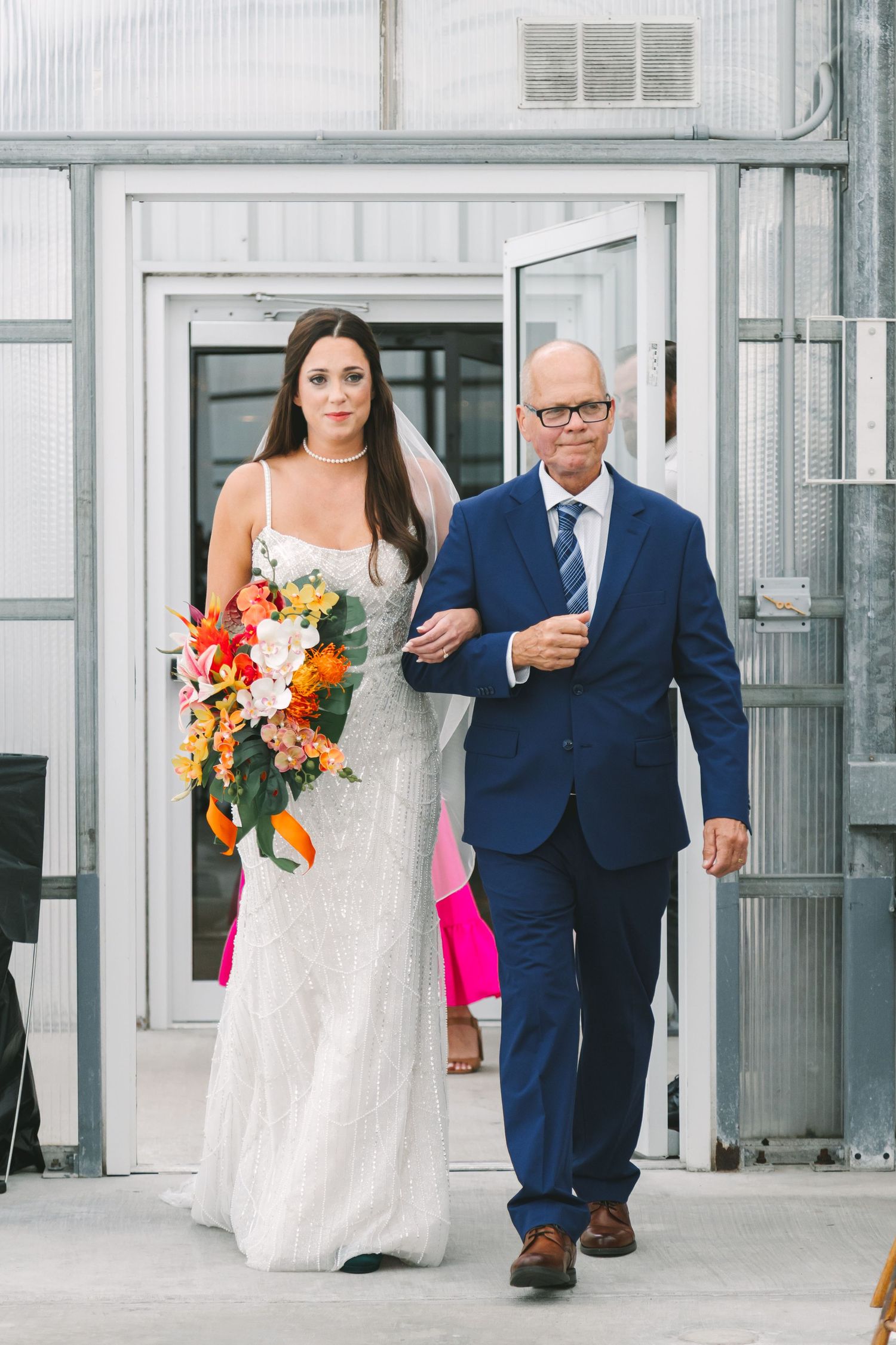 Bride in white lace gown holding colorful bouquet walks down aisle with father in navy suit.