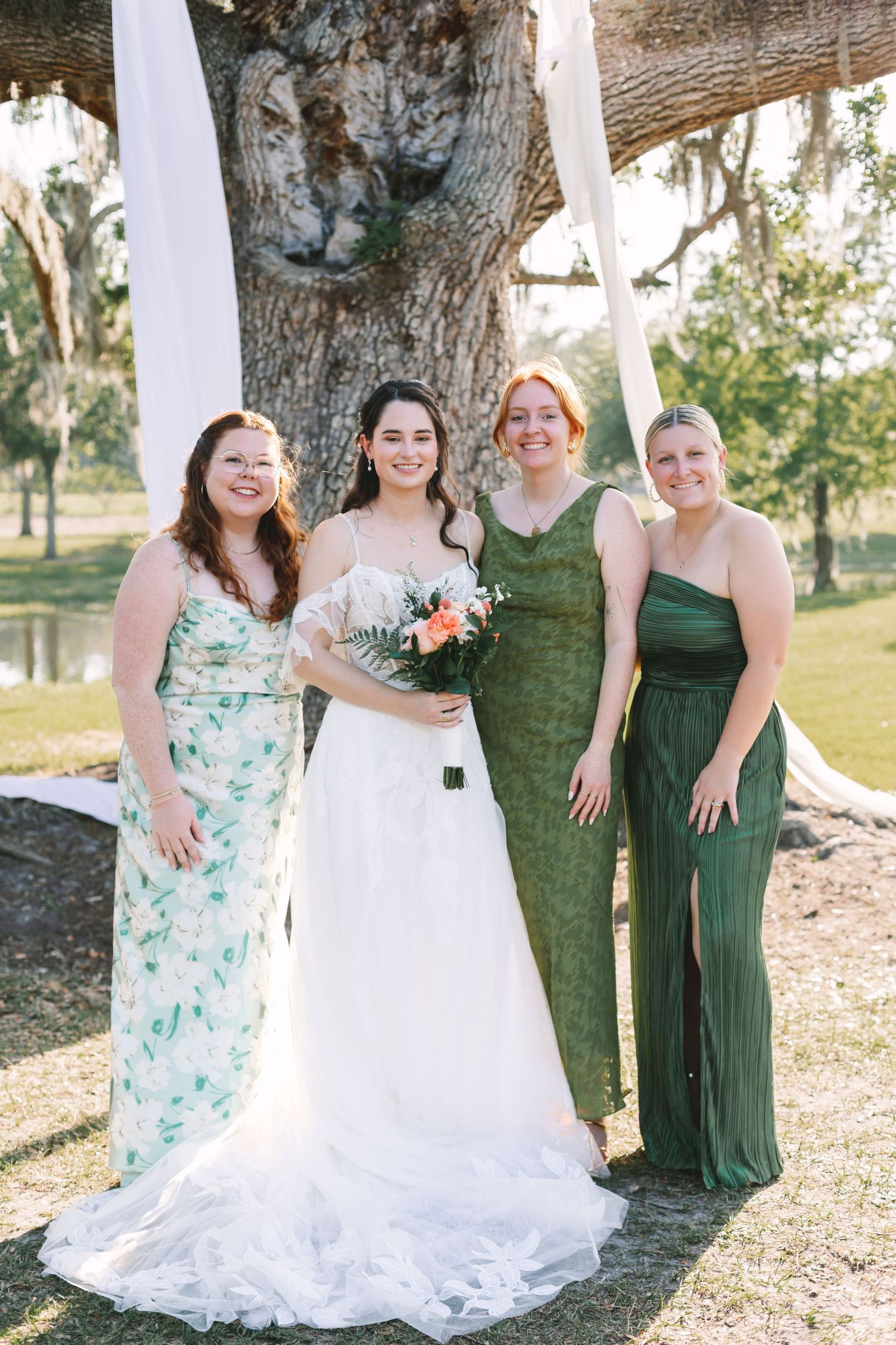 Bride with bridesmaids in green dresses posing under large oak tree at outdoor wedding.