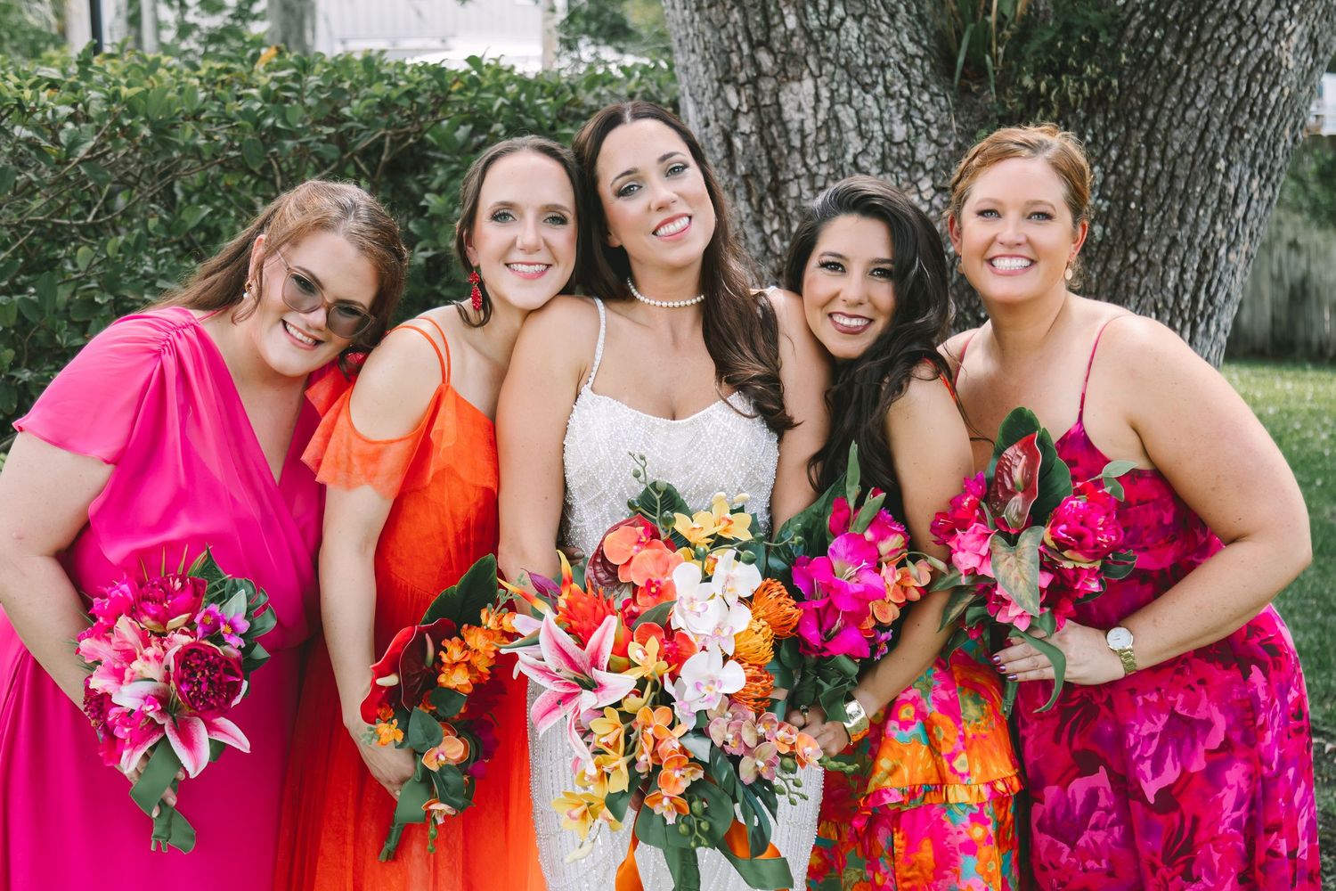 Bride in white gown with four bridesmaids in vibrant pink and orange dresses holding tropical floral bouquets outdoors.