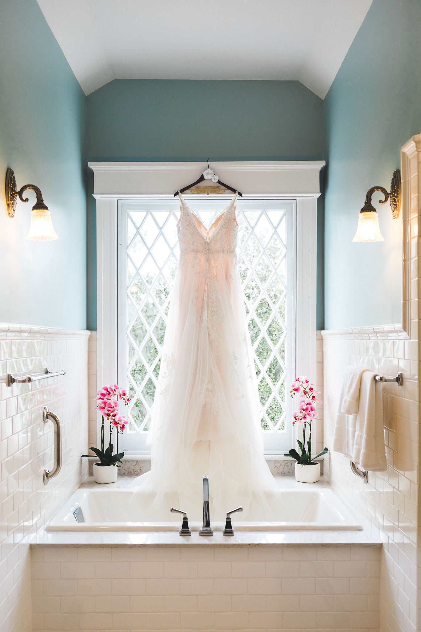 Elegant wedding gown hanging on diamond-paned window above bathtub with pink orchids.