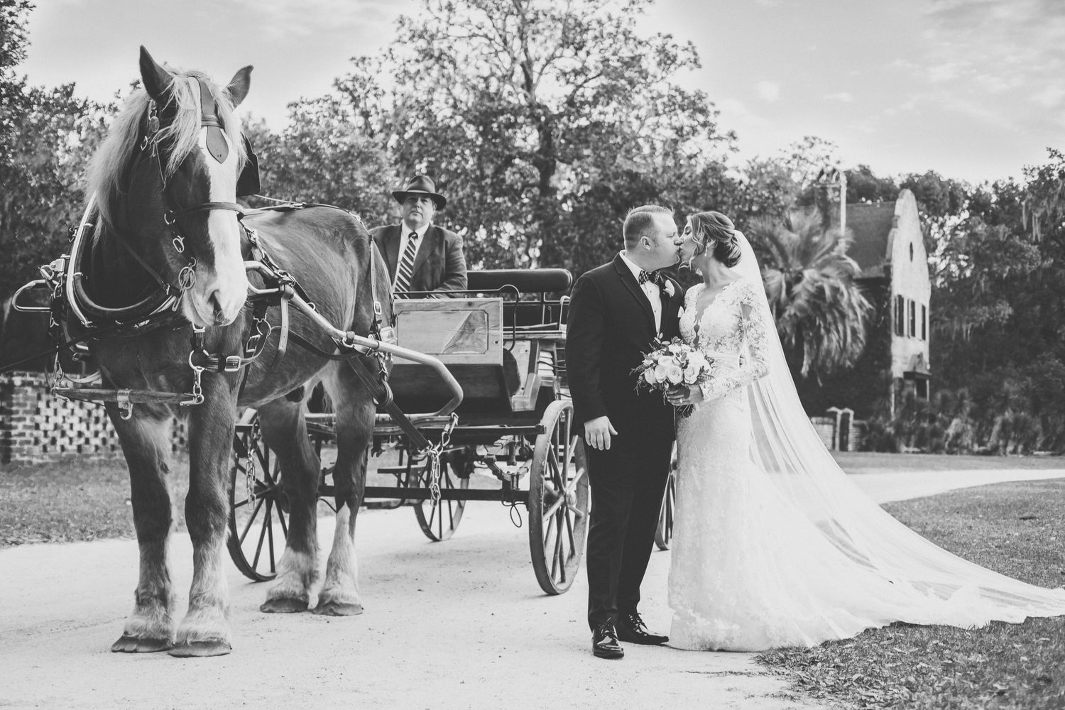 Black and white wedding photo of bride and groom kissing beside a horse-drawn carriage outdoors.