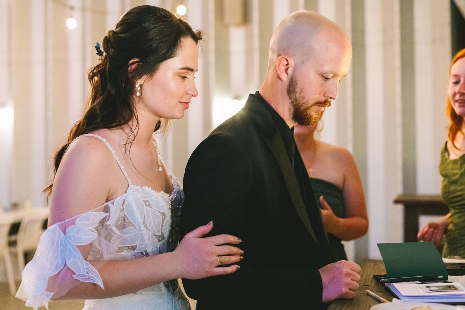 Bride in off-shoulder white gown and bearded groom in black suit review documents at wedding signing.