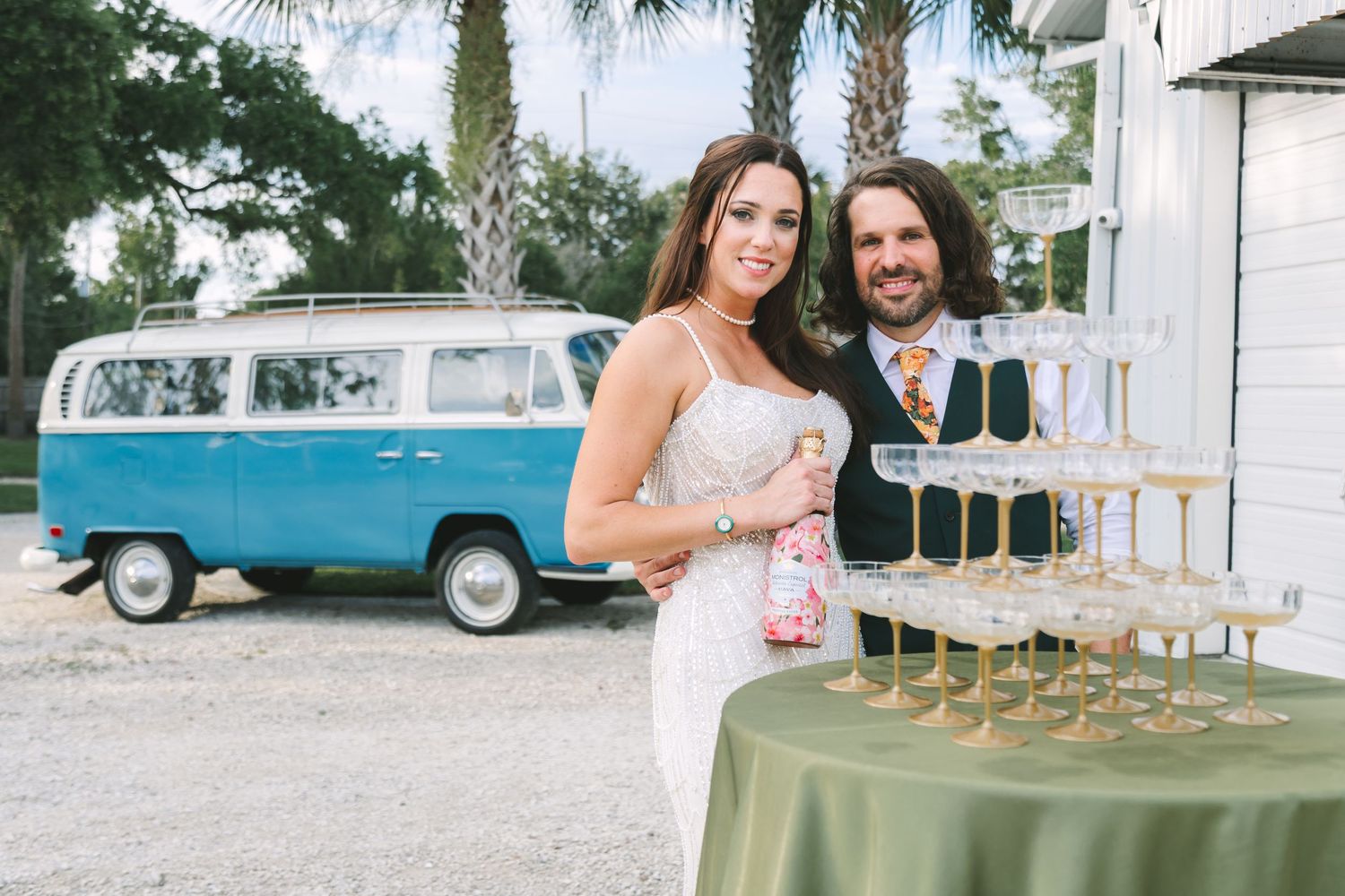 Bride and groom posing beside champagne tower with vintage blue VW bus in background.