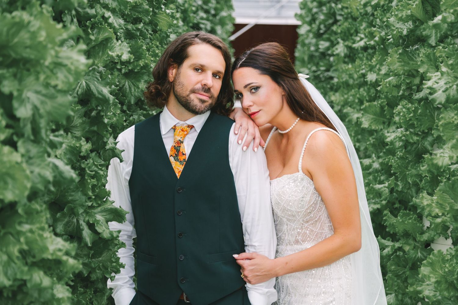 Bride and groom pose romantically among lush green hedges, groom in dark suit with floral tie.