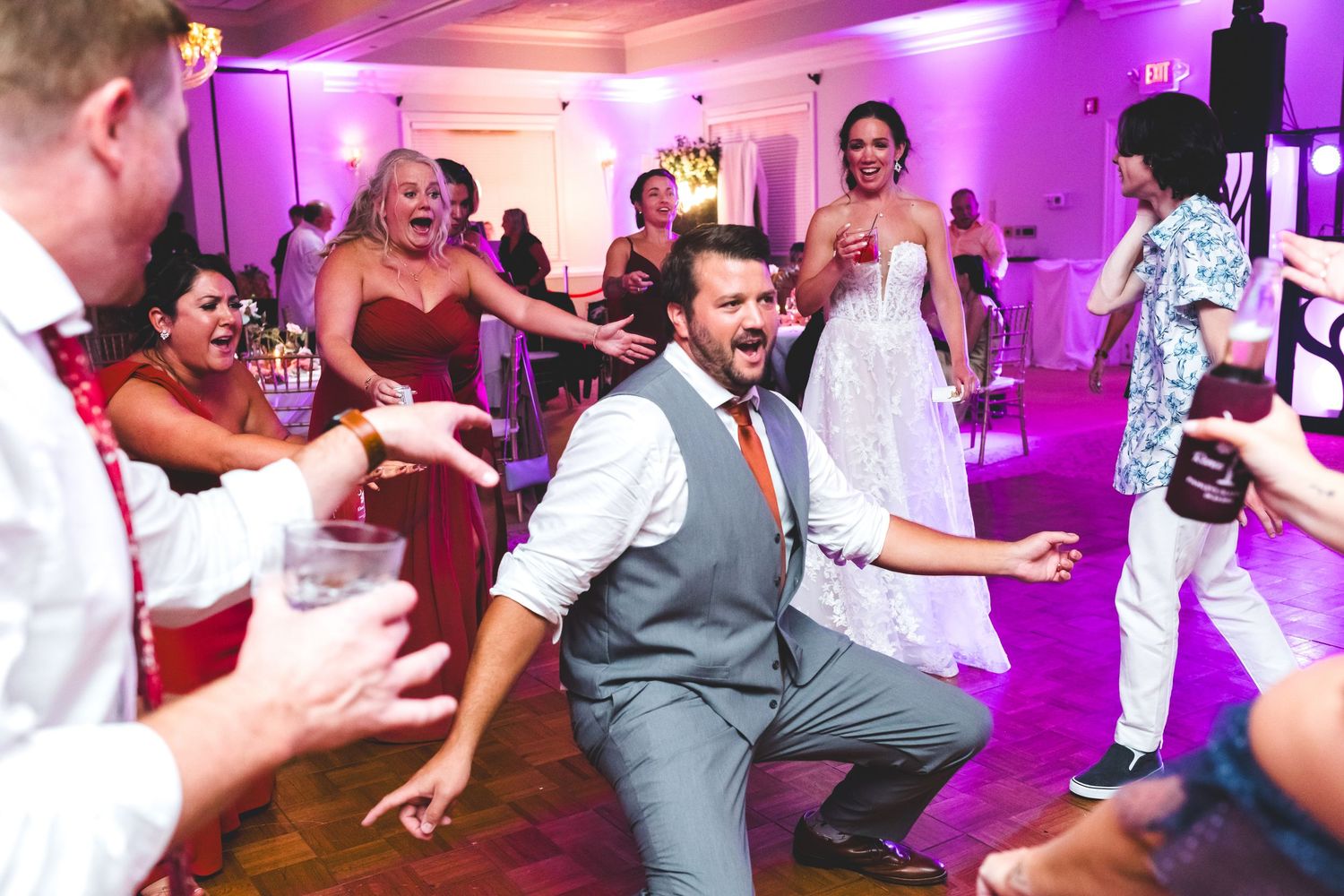 Energetic wedding reception dance floor with groom in gray suit dancing while guests cheer under purple lights.