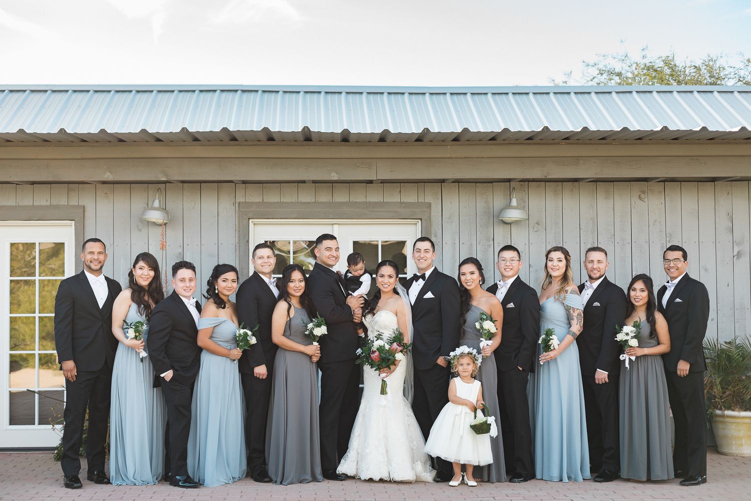 Wedding party poses in dusty blue bridesmaid dresses and gray suits outside barn venue.