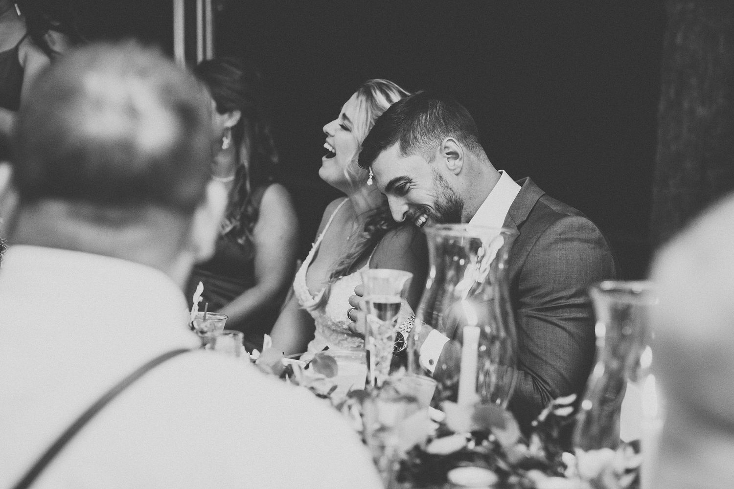 Black and white photo of wedding guests laughing and sharing joyful moments at the reception dinner table.